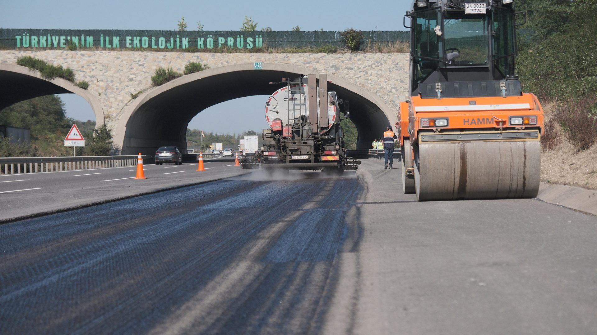 Road work with a spraying truck, roller, and an ecological bridge overhead.