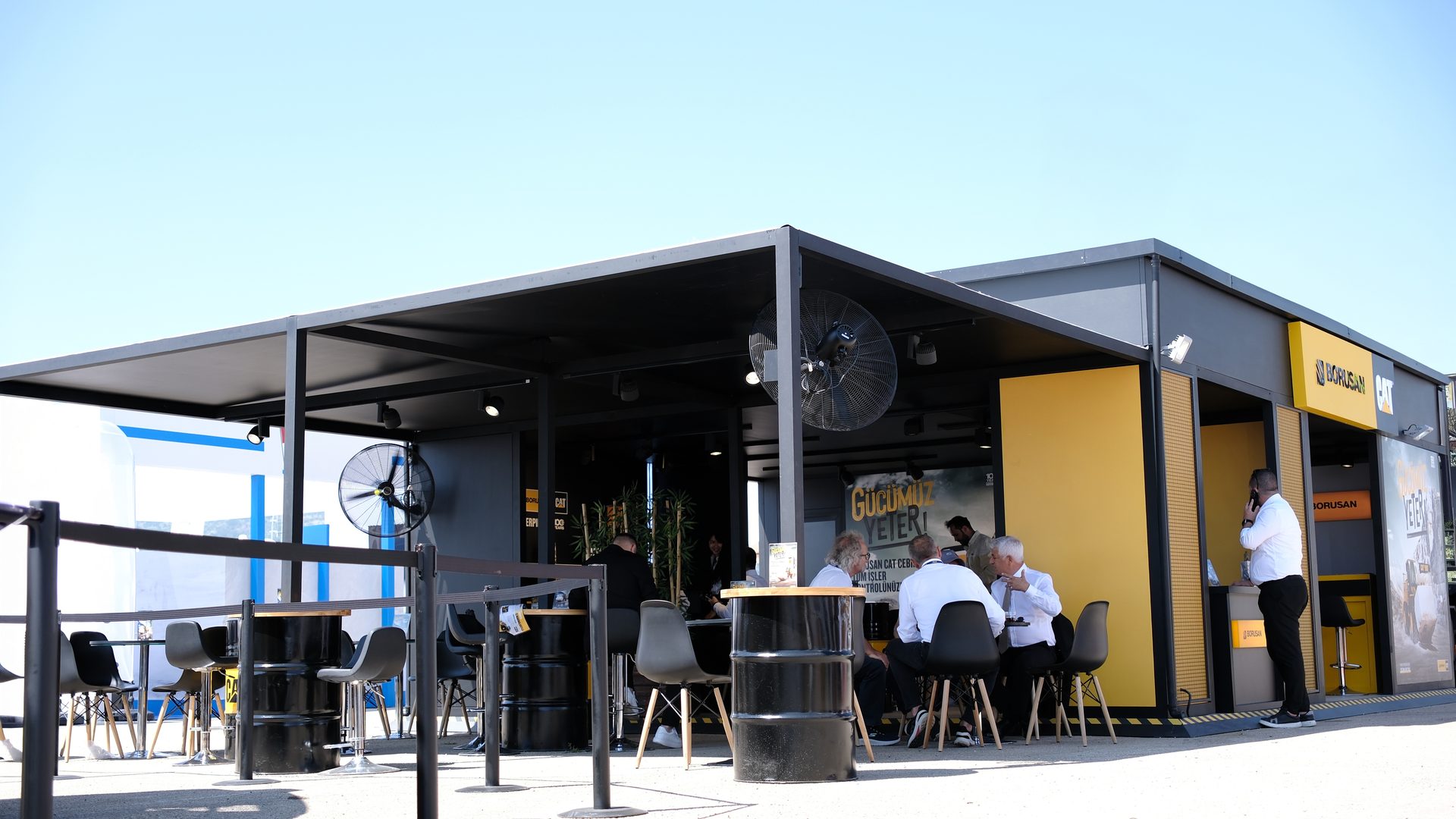 Outdoor Borusan CAT booth with people gathering around barrel tables, under fans.