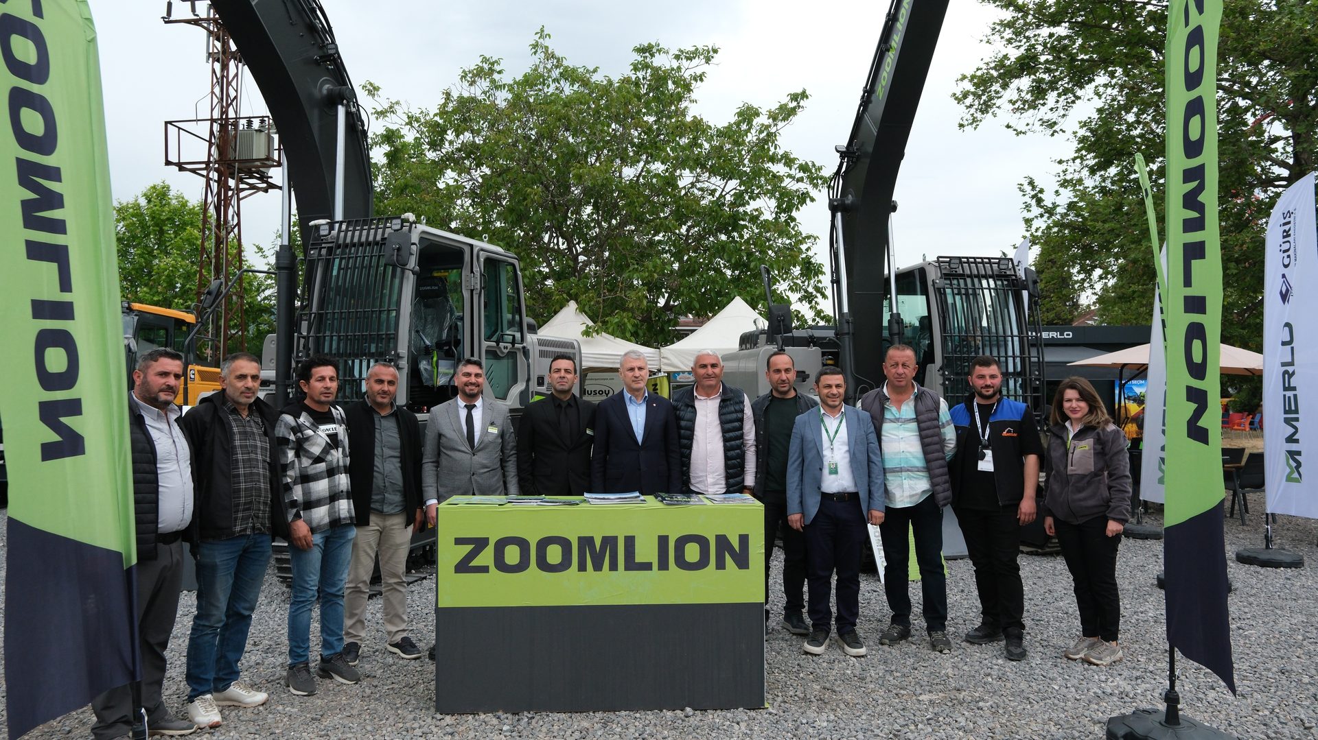 A group of people stand in front of two large excavators and a 'ZOOMILION' sign at an outdoor event.