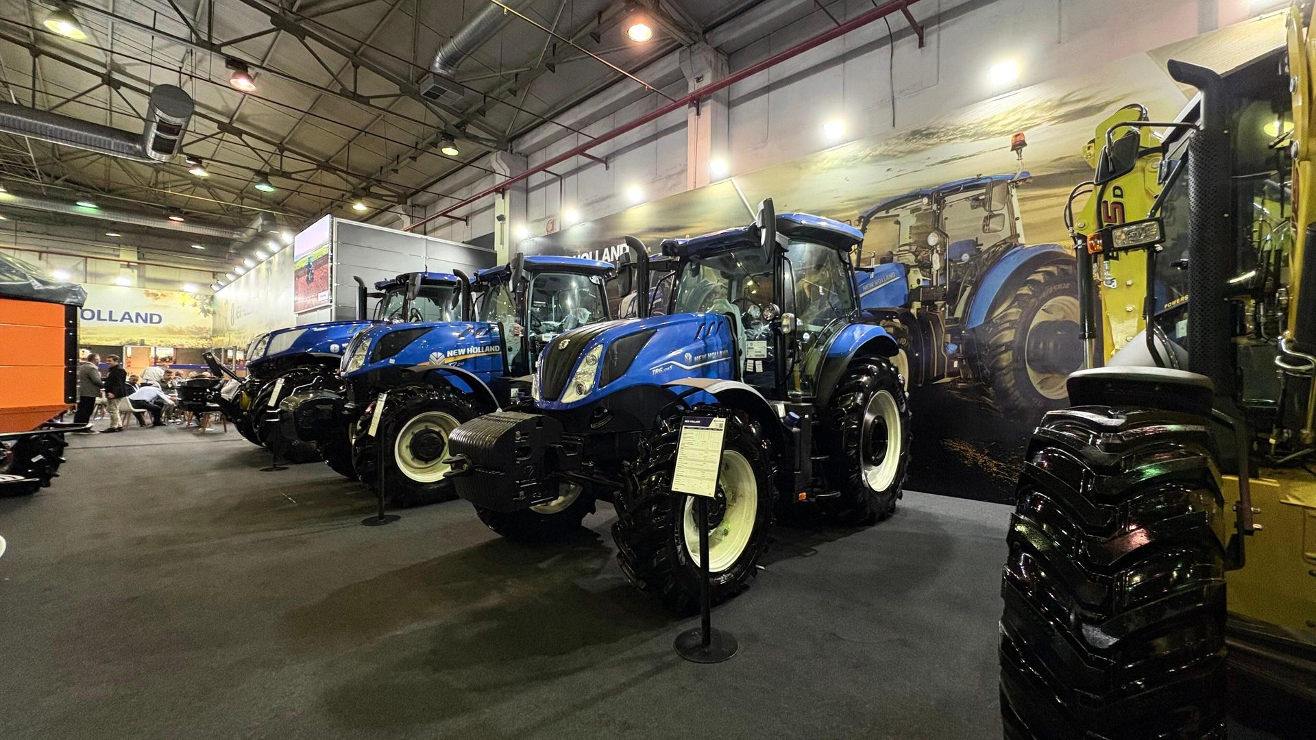 Blue New Holland tractors lined up at an indoor exhibition with a brand banner.