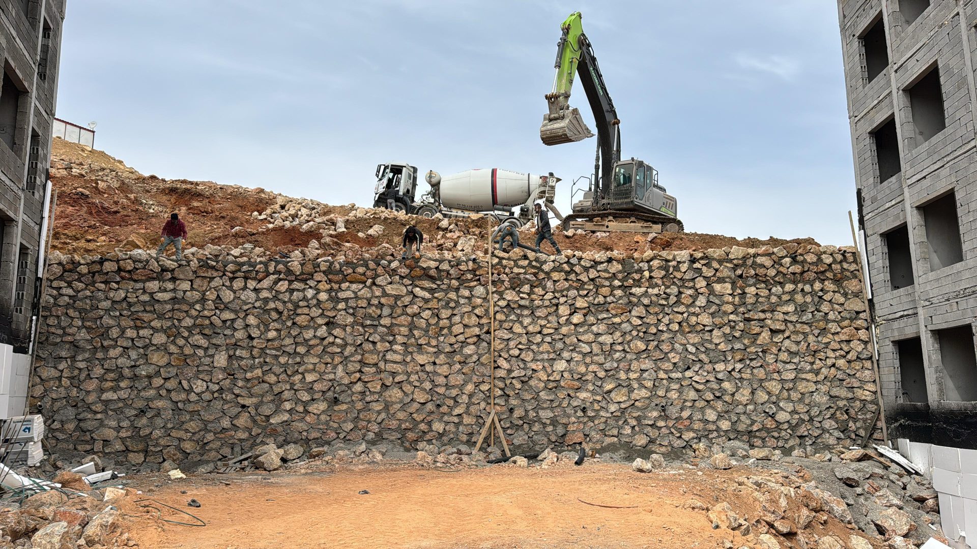 Construction site with a large stone retaining wall, excavator, cement truck, and workers on a hill.