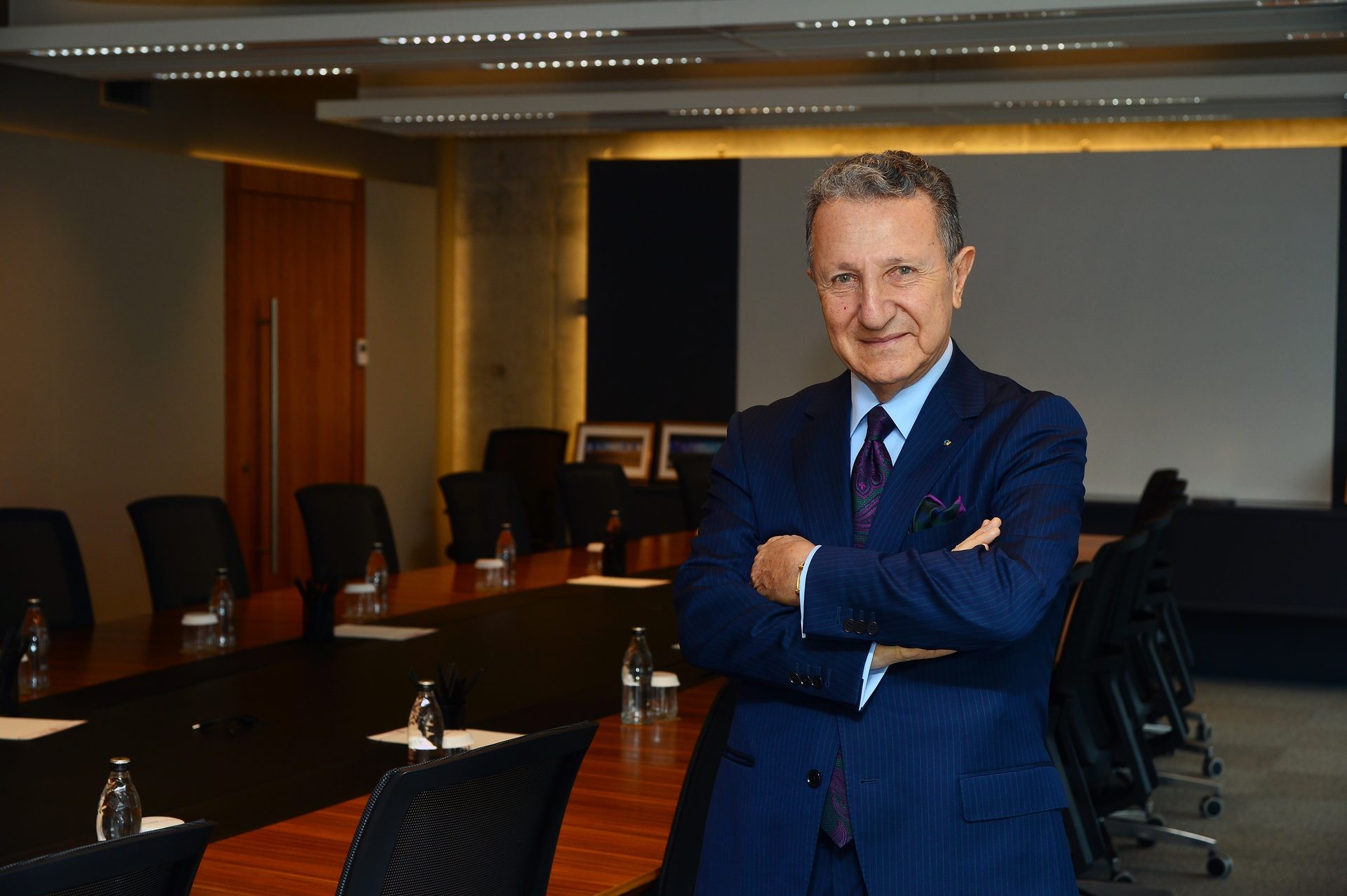 A smiling man in a blue pinstripe suit with arms crossed in a modern boardroom setting.