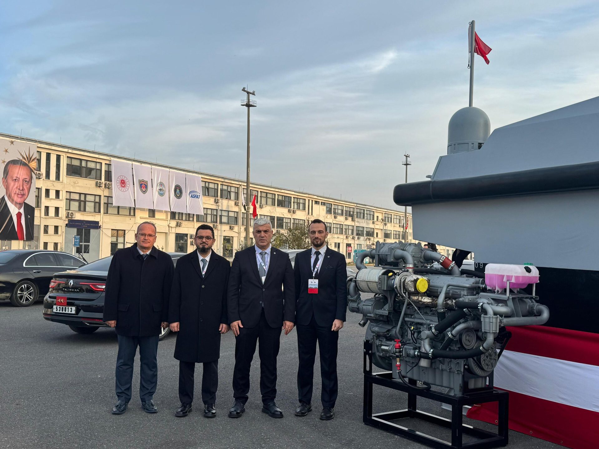 Four men stand with a marine engine and a boat; a building and banners with Erdoğan's portrait are behind them.