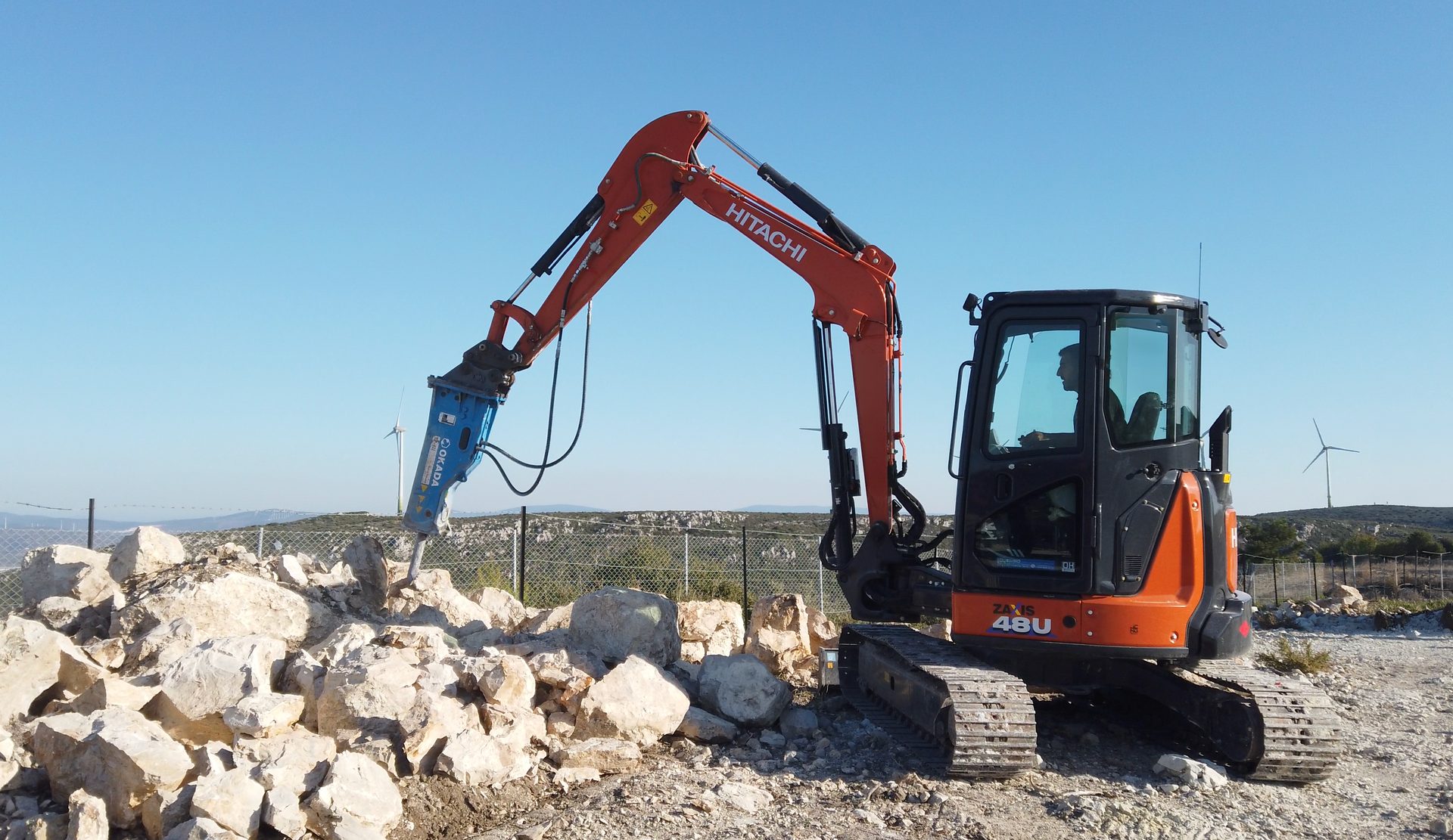 An orange Hitachi mini excavator with a hydraulic breaker attachment breaking rocks under a clear blue sky.