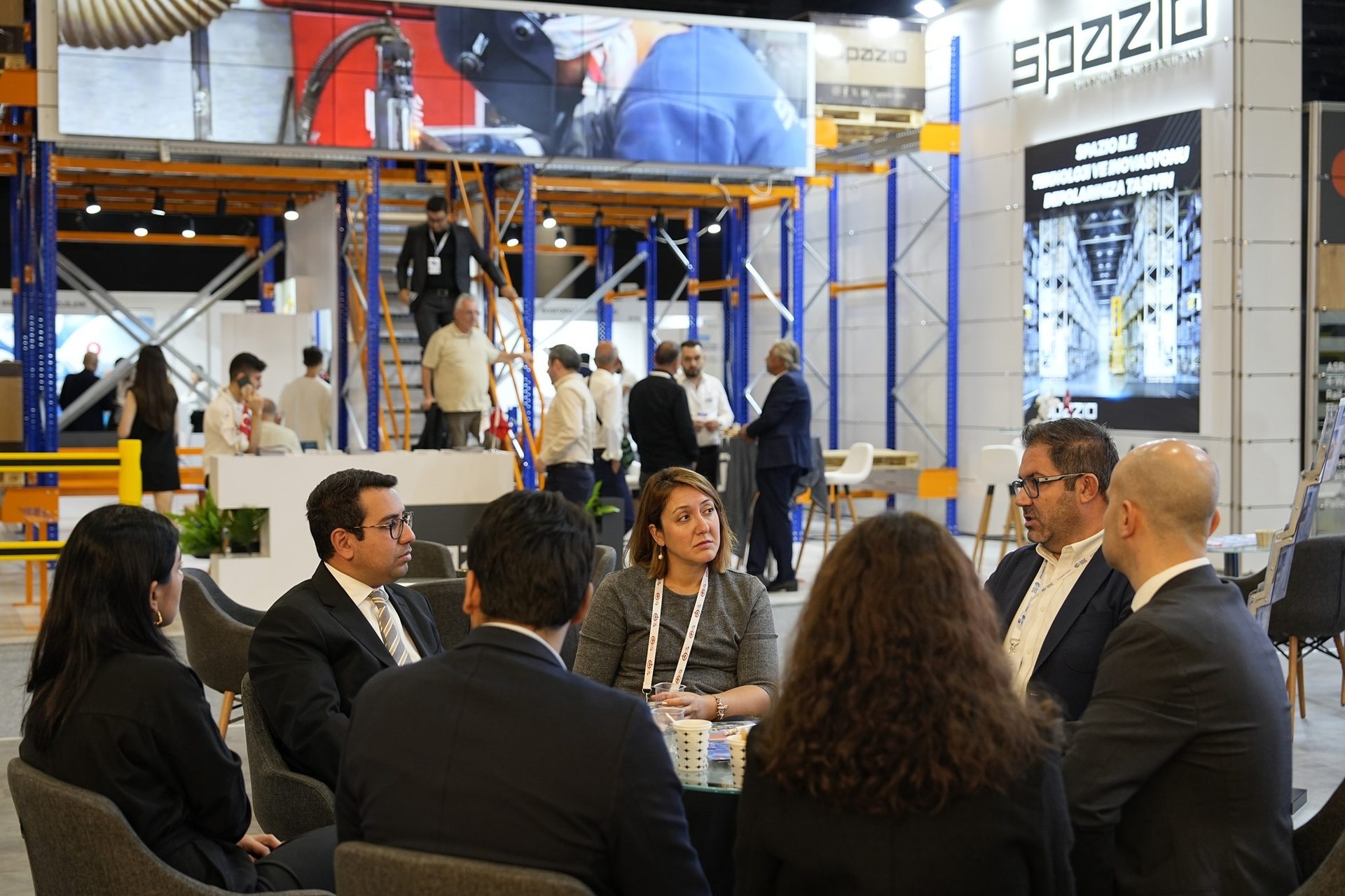 Group of professionals discussing at a Spazzio trade show booth with industrial shelving in the background.
