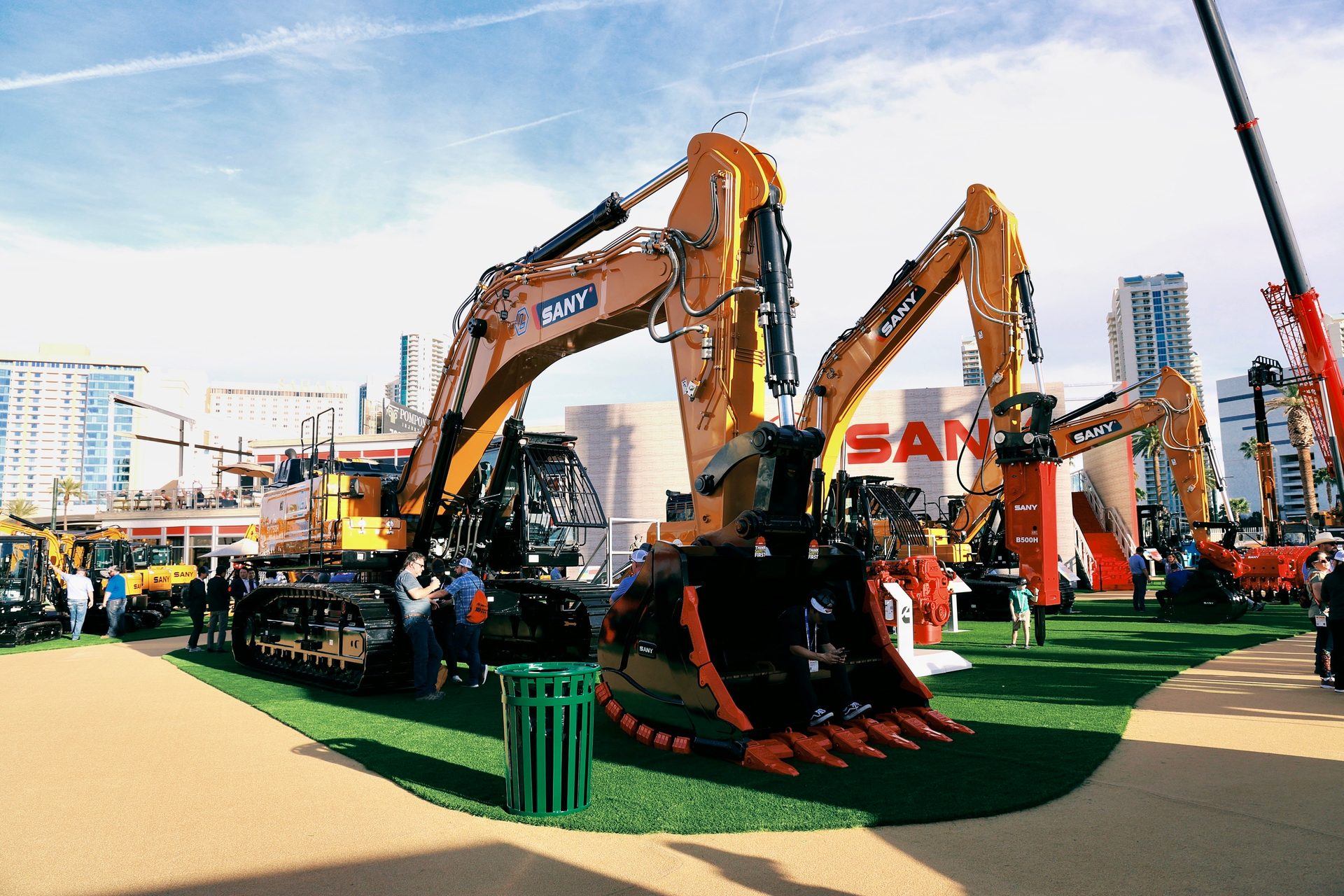 Large SANY excavators on display at an outdoor exhibition with people, city buildings, and a crane.