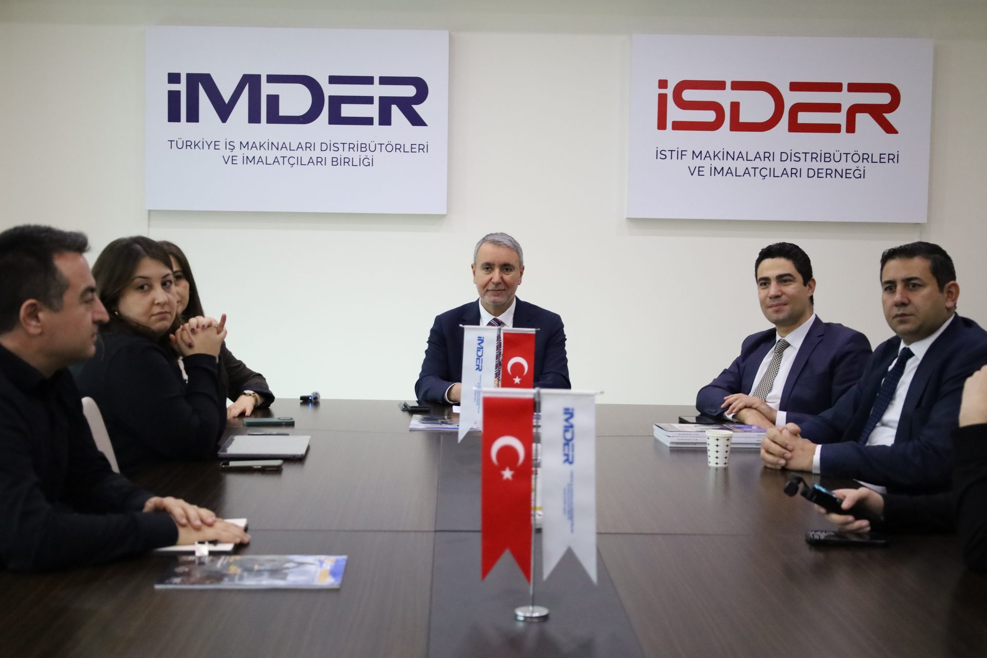 People at a business meeting with IMDER and ISDER logos on the wall, and Turkish flags on the table.