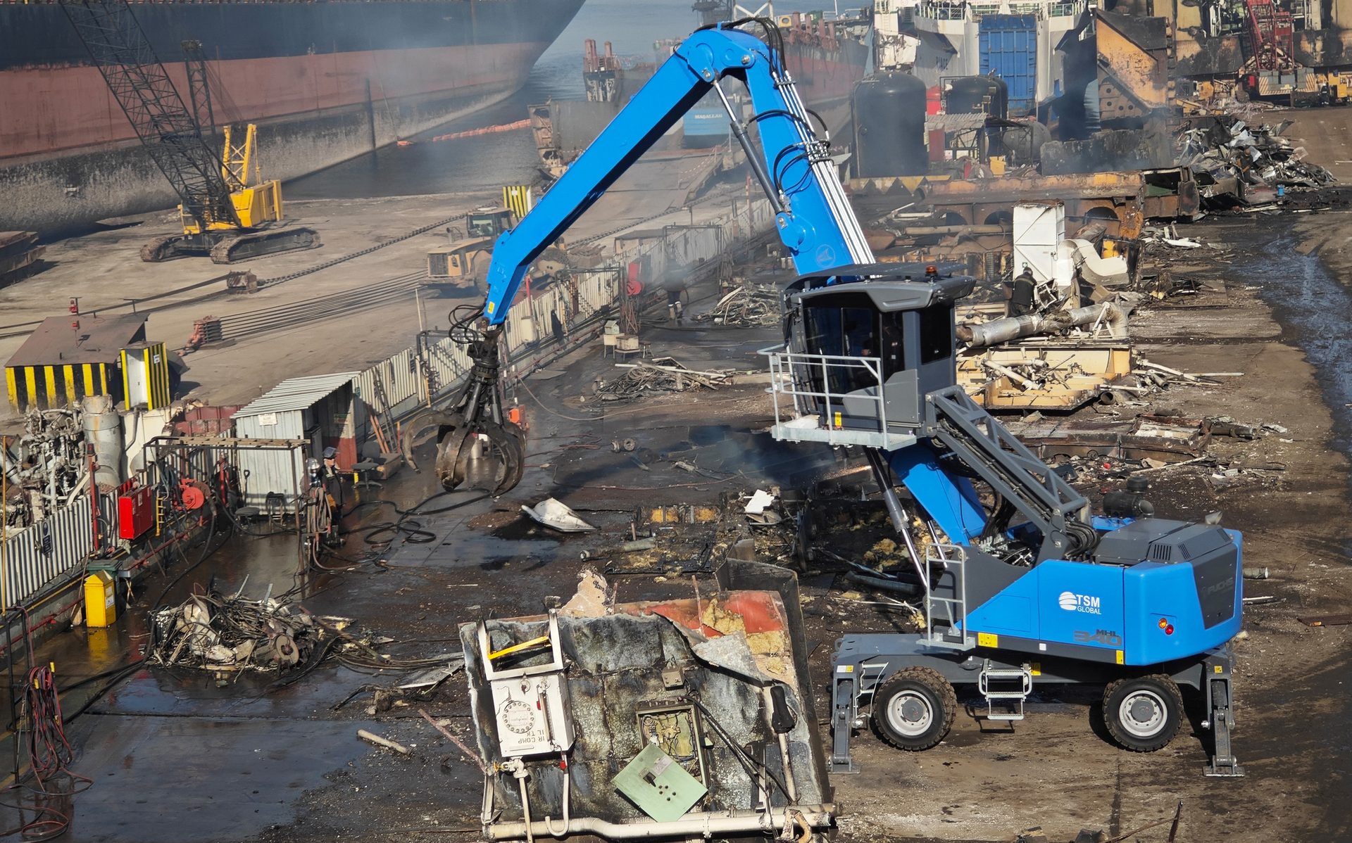 A blue material handler with a grapple processes scrap metal in a busy shipyard with a large ship.