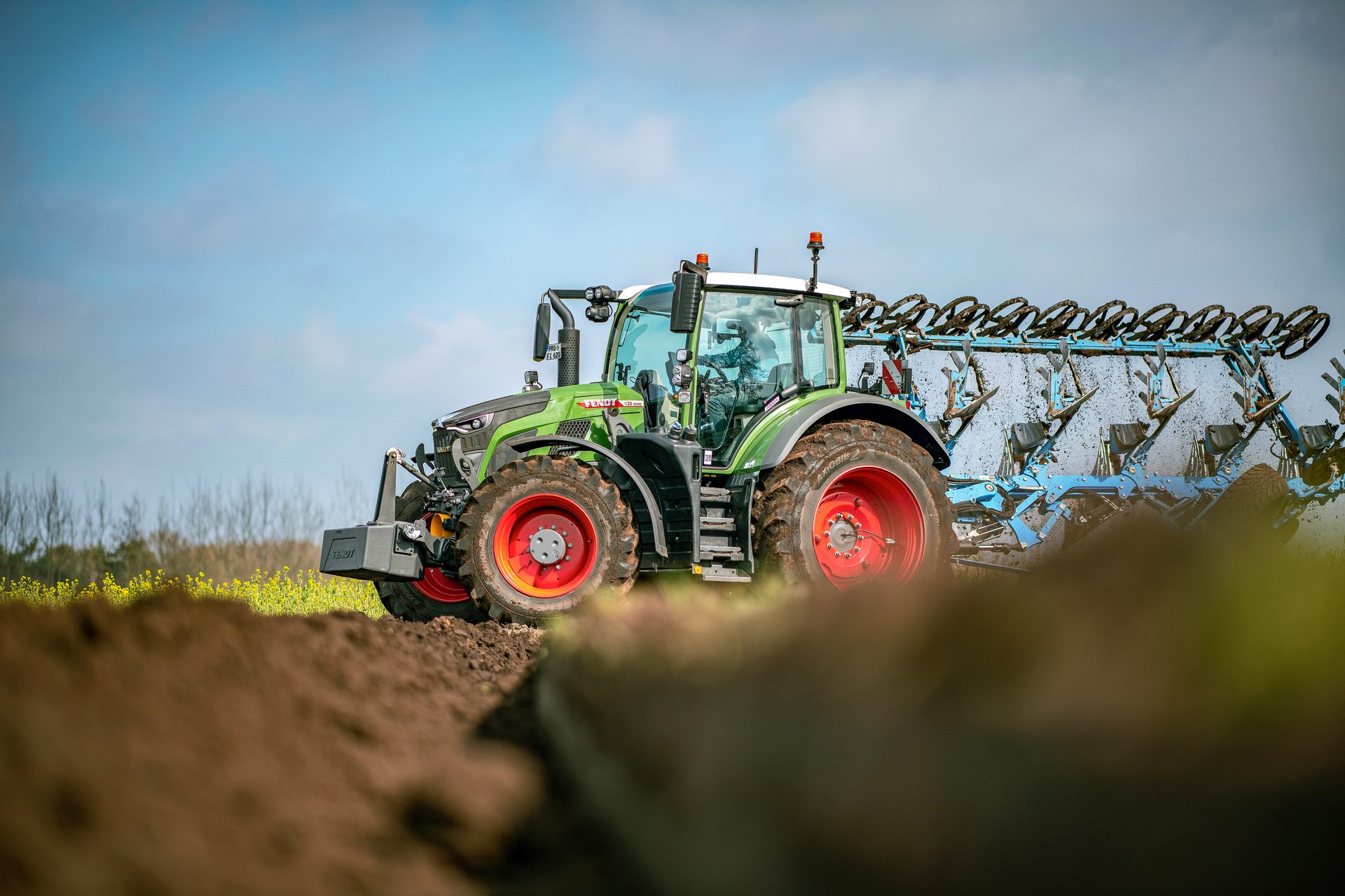 A green Fendt tractor plowing a field with a large blue plow attachment under a blue sky.