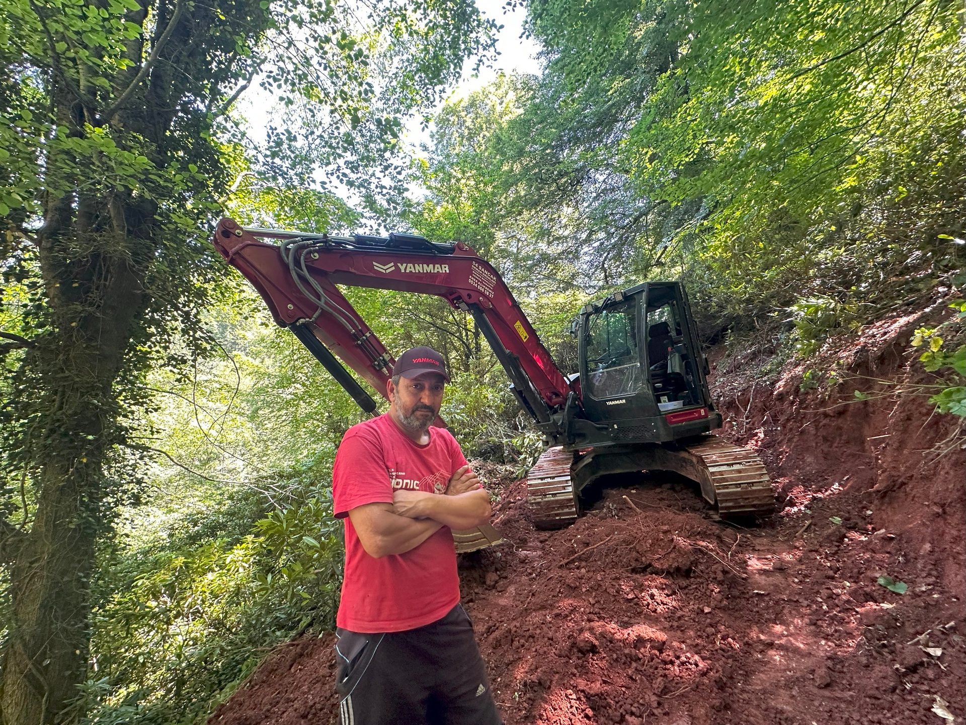 Man with crossed arms by a red Yanmar excavator on a red dirt path in a green forest.