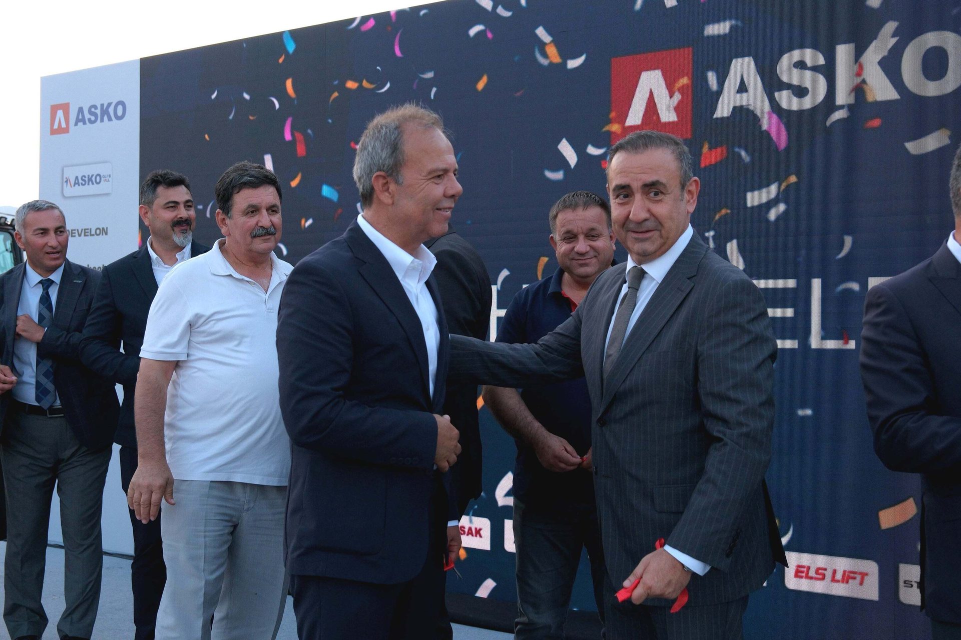Two men in suits shake hands in front of a large screen with the "ASKO" logo and confetti.