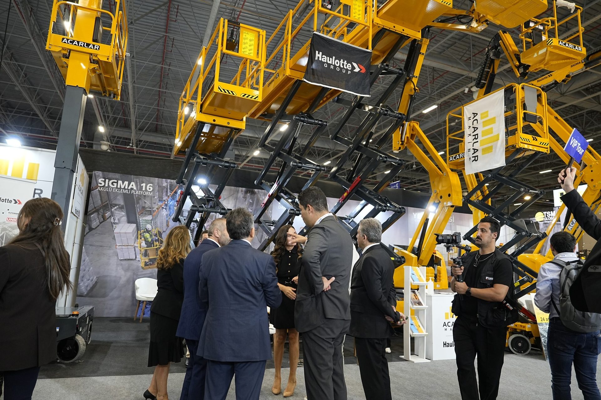 People view yellow Acarlar and Haulotte lifting platforms at a trade show.