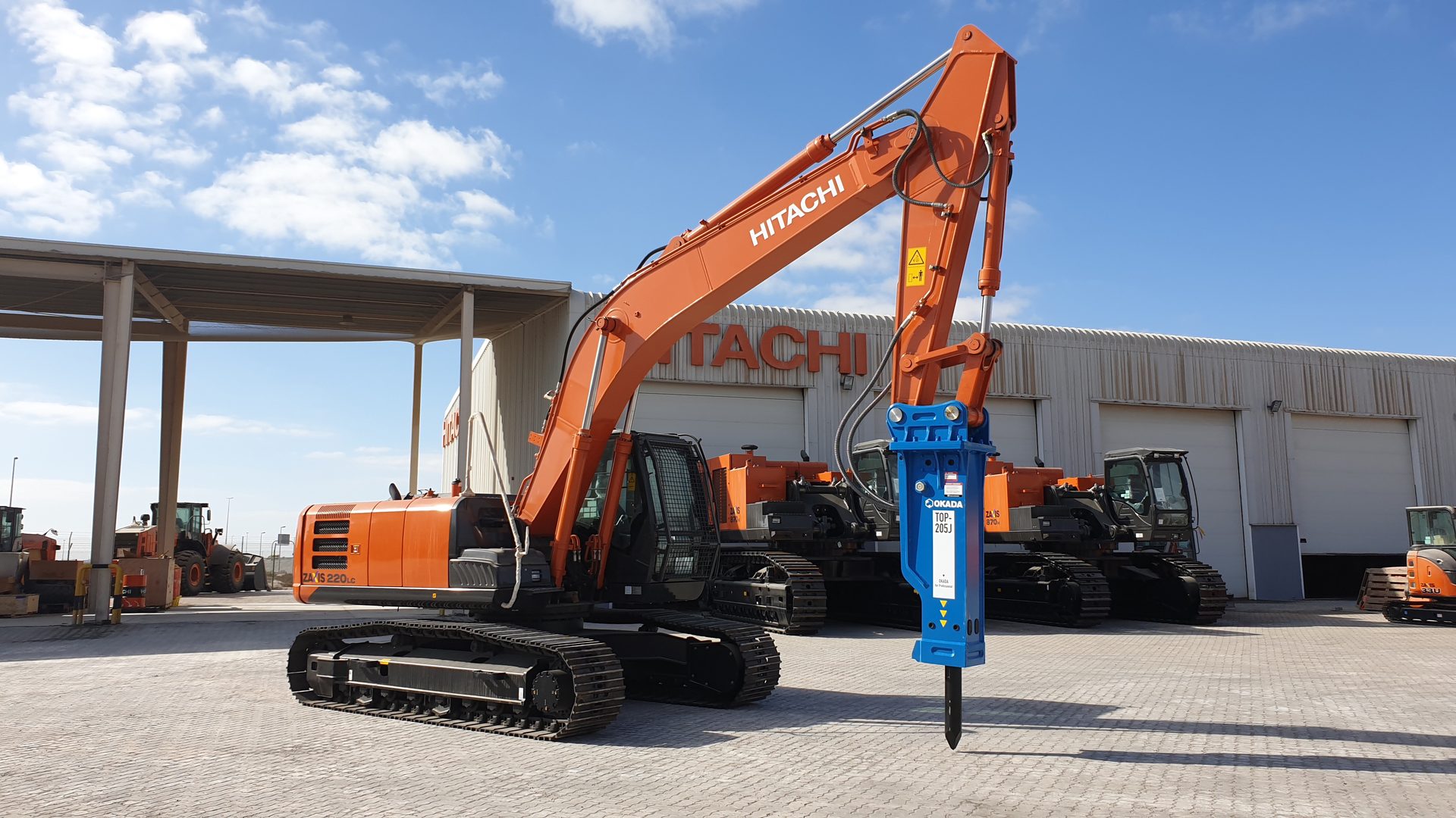 An orange Hitachi excavator with a blue Okada hydraulic breaker attachment in a construction yard.