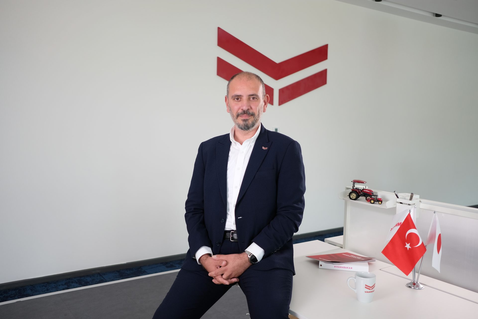 Man in a suit sitting on a desk in an office with a red logo, flags, and a miniature tractor.