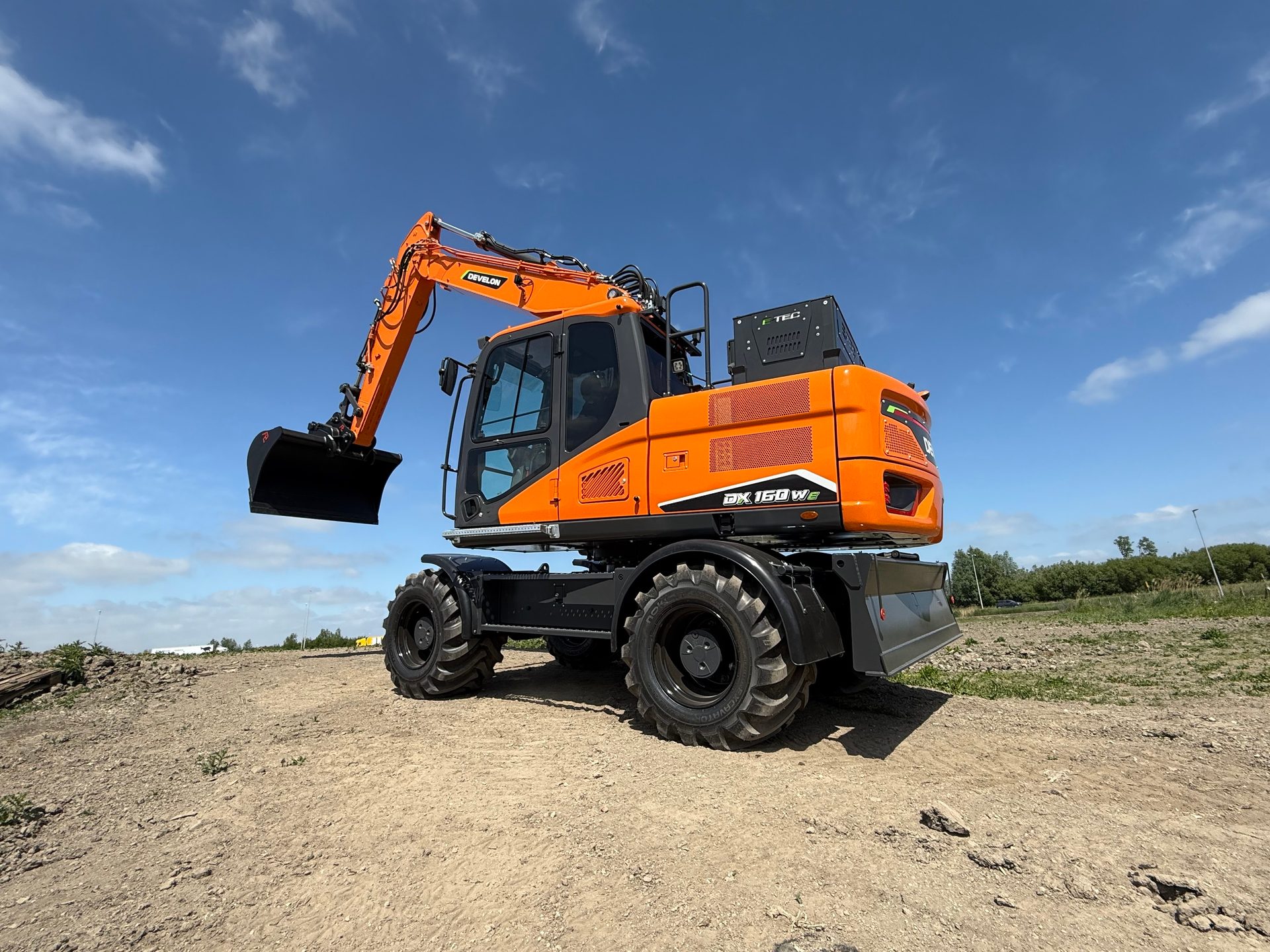 Orange Develon wheeled excavator on dirt under blue sky.