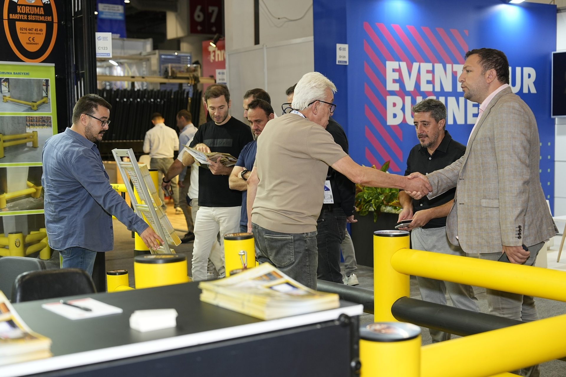 Men shaking hands at a trade show booth with yellow barriers and an "EVENT YOUR BUSINESS" sign.