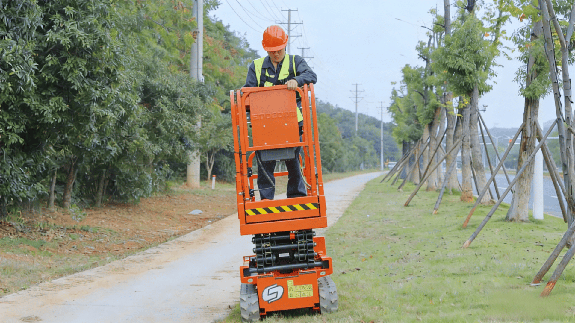 A worker in safety gear operates an orange scissor lift on a paved path surrounded by trees.