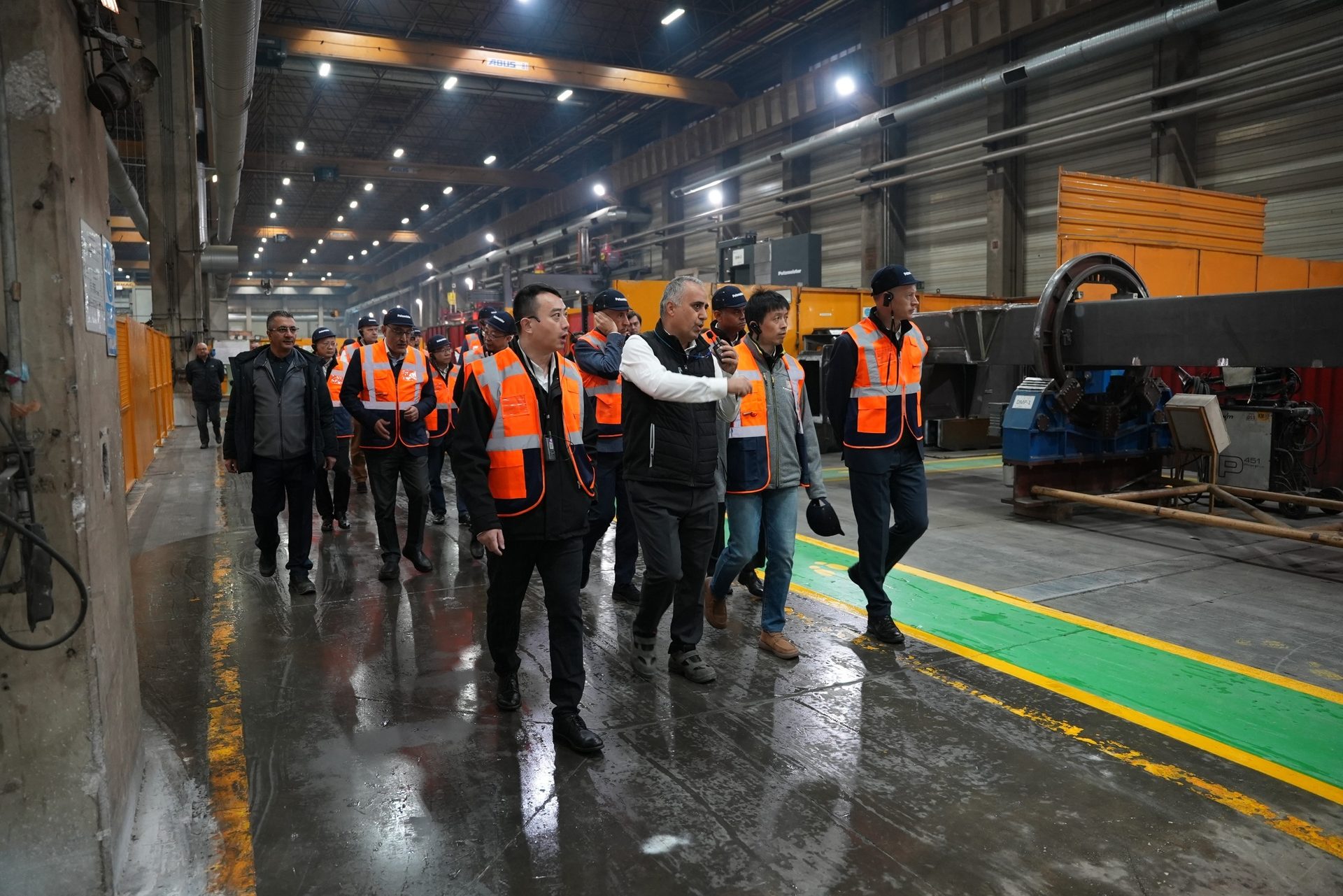 A group of people in orange safety vests walking through a factory, one speaking and gesturing.