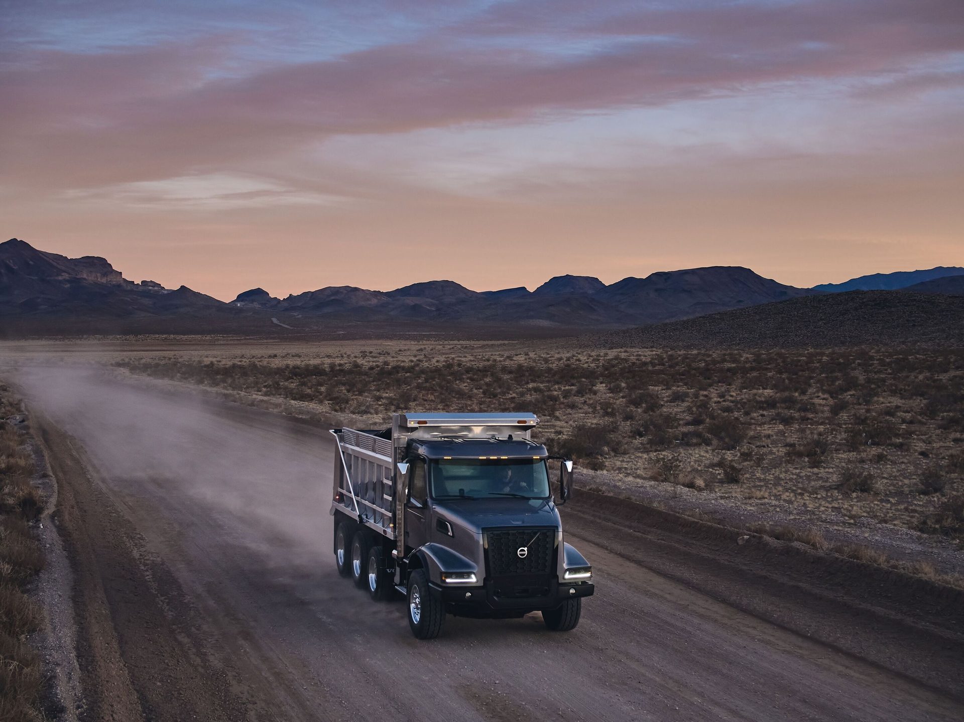 A dark gray Volvo dump truck drives on a dusty dirt road through a vast desert landscape at sunset.