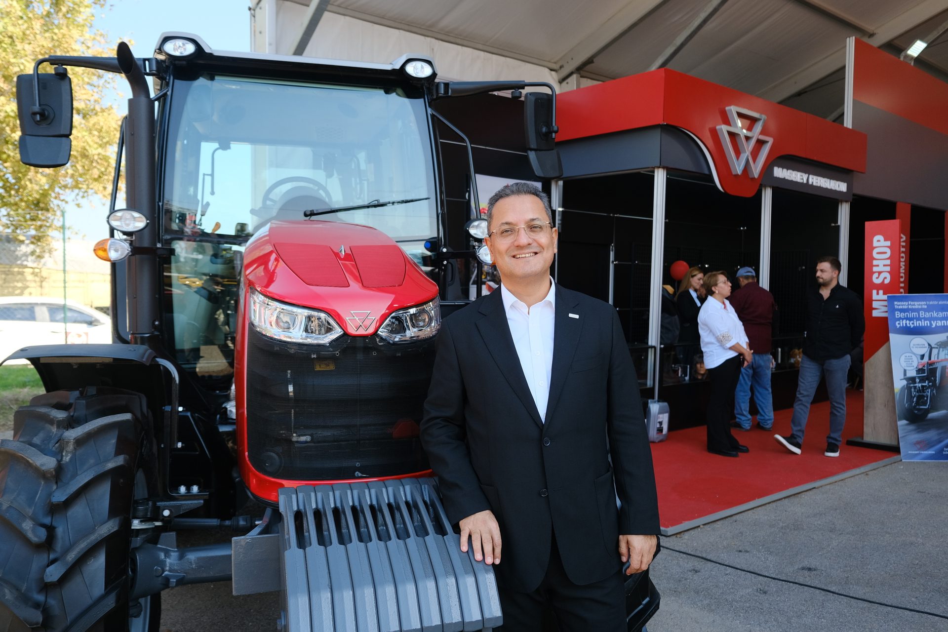 A man in a suit smiles next to a red Massey Ferguson tractor and booth at an outdoor event.