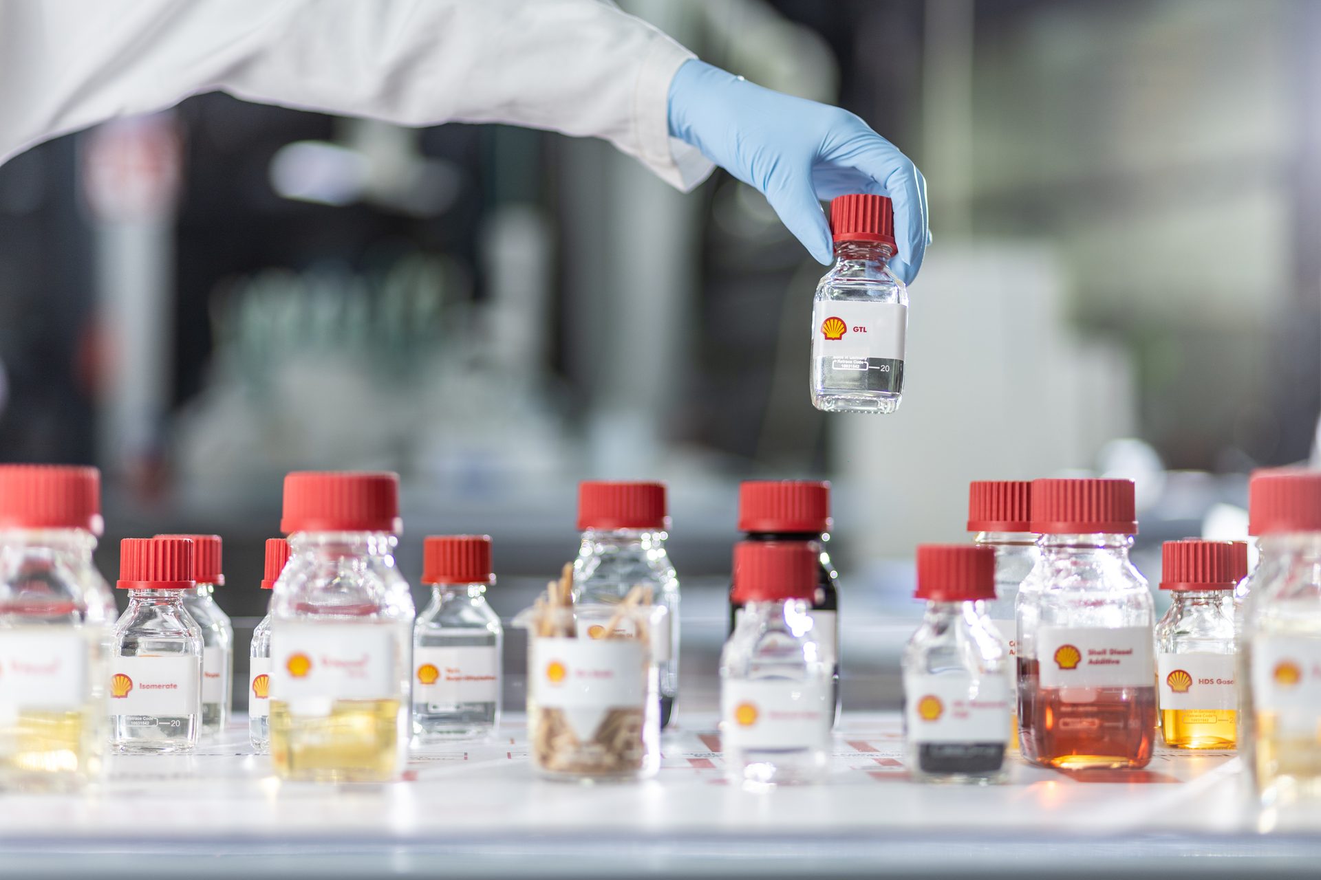 A researcher's gloved hand holds a Shell GTL sample bottle, with more samples on a lab table.