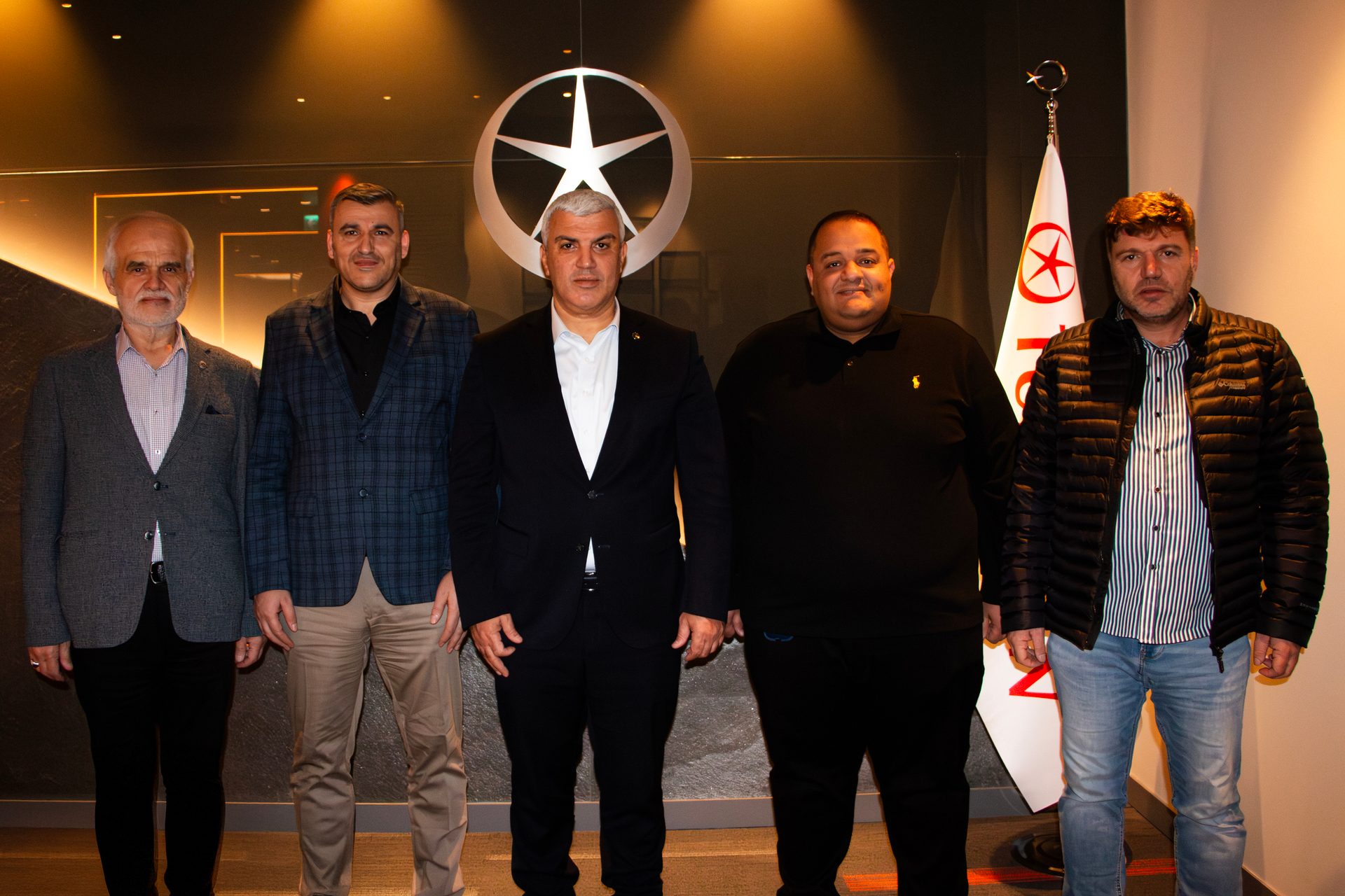 Five men pose indoors in front of a wall featuring a star logo and a flag.