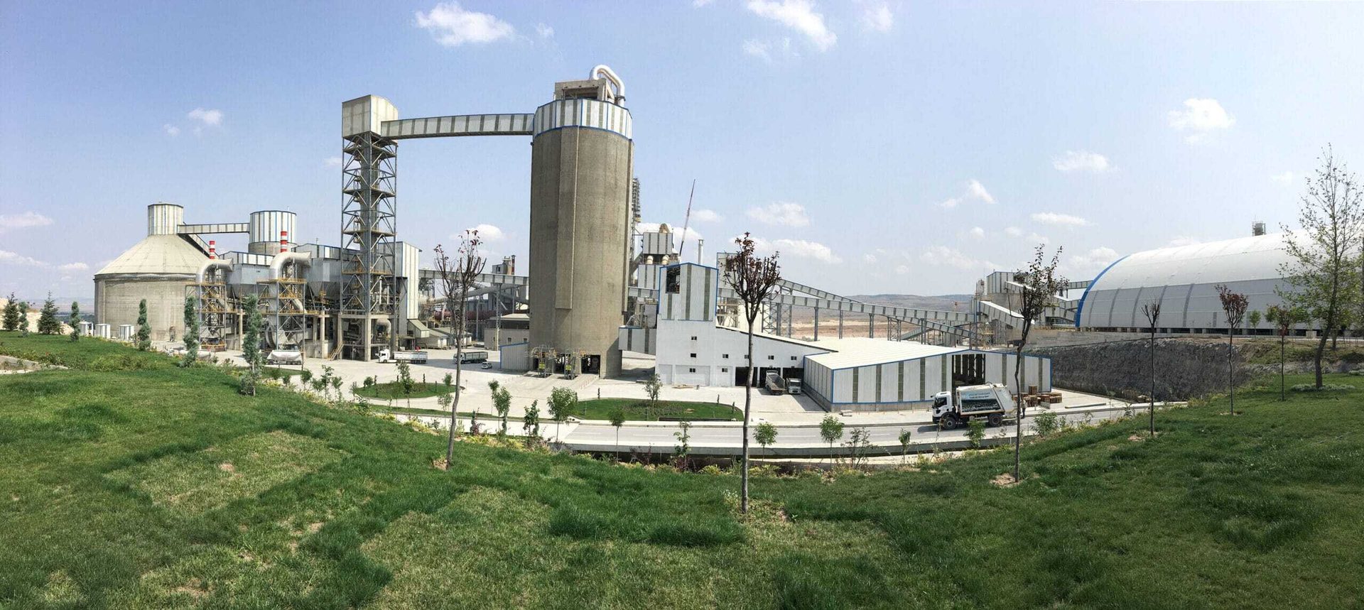 Panoramic view of a large industrial plant with silos, conveyor belts, buildings, and green grass.
