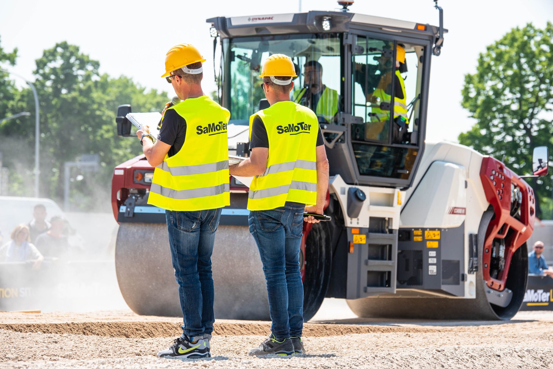 Two construction workers in safety gear stand by a large road roller.
