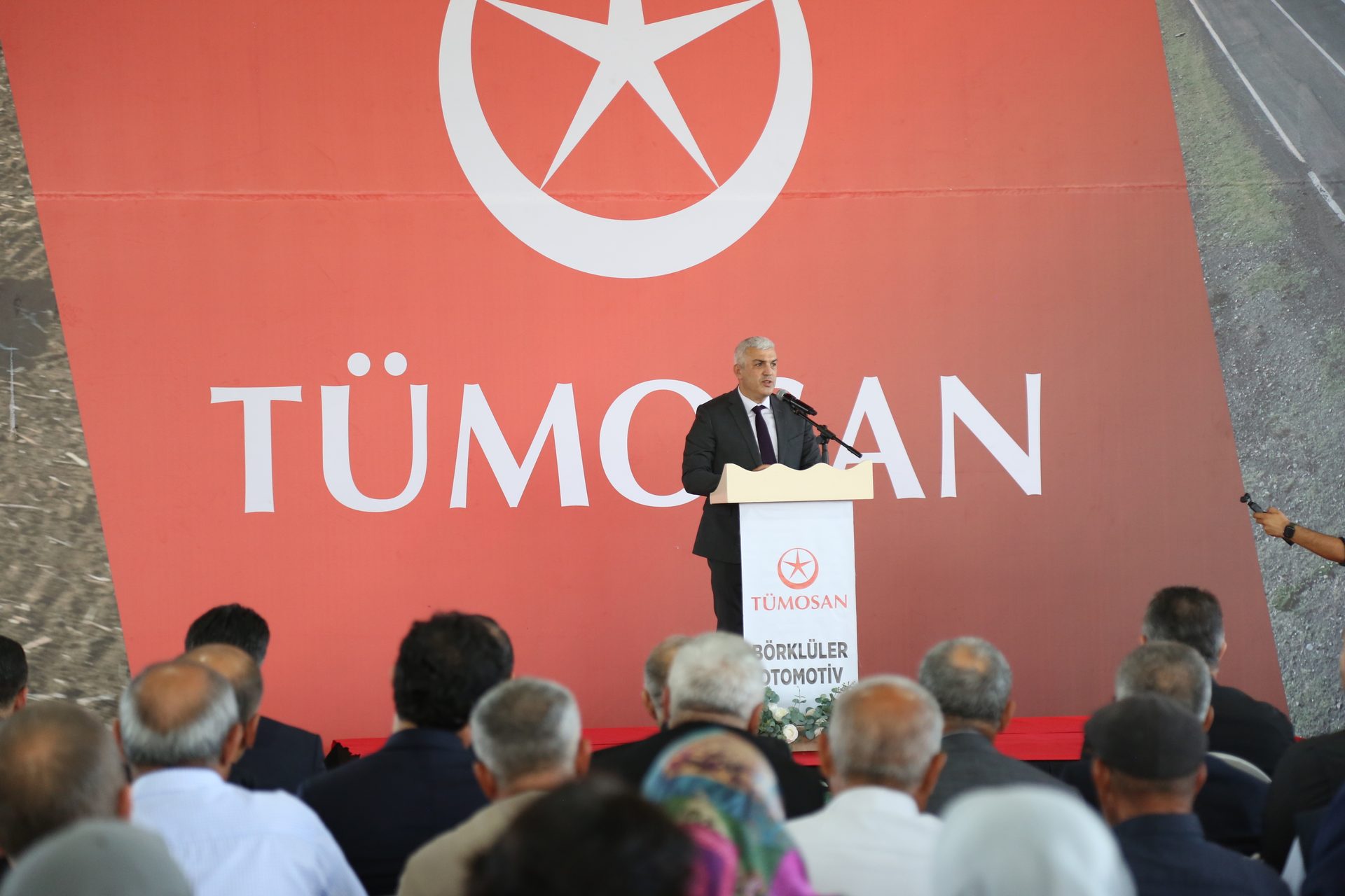 Man speaking at a podium in front of a large red TÜMOSAN banner with an audience.