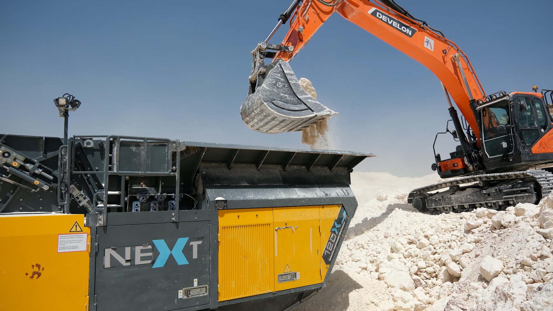 Orange excavator loads crushed rock into a NEXT mobile crusher.
