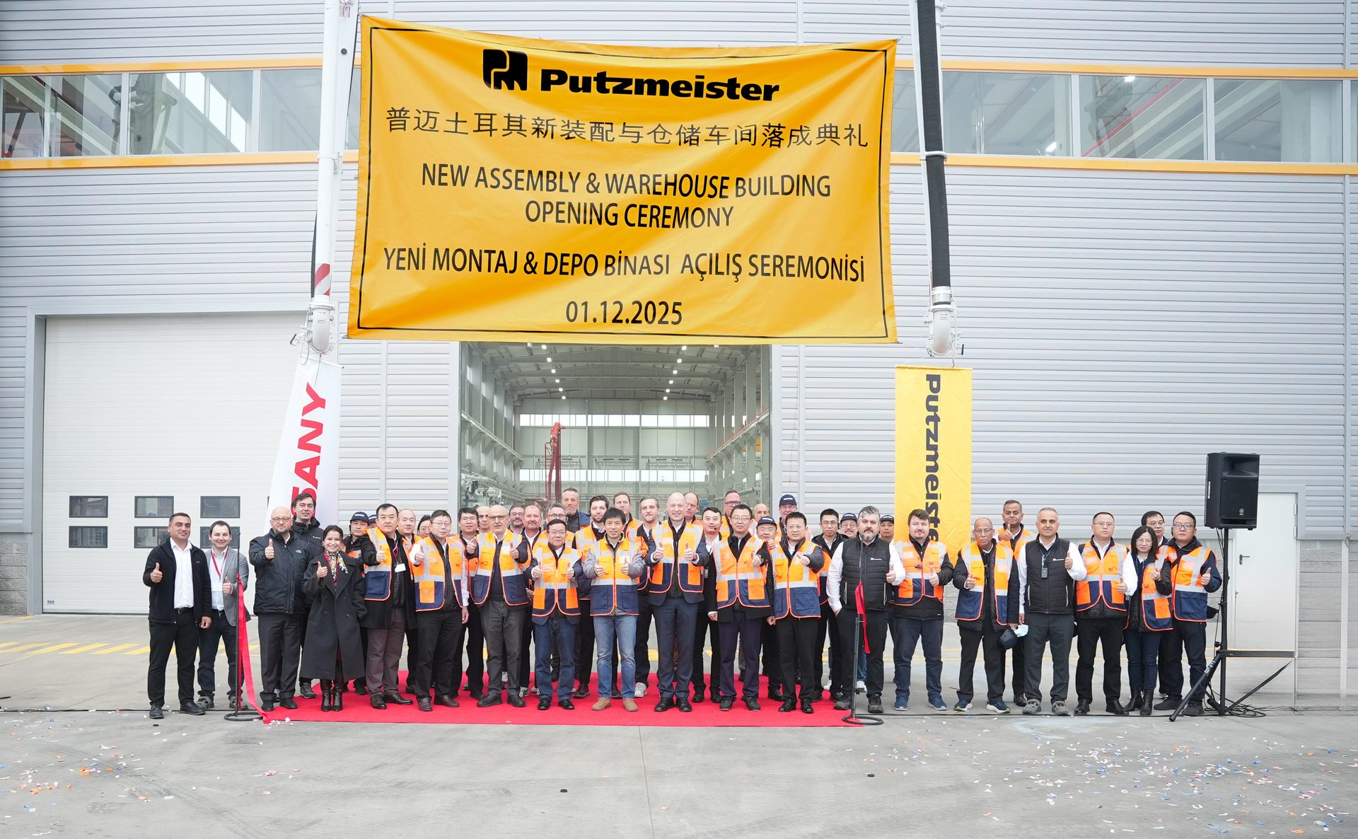 A group of people in safety vests stand on a red carpet in front of a building with a "Putzmeister New Assembly & Warehouse Building Opening Ceremony" banner dated 01.12.2025.