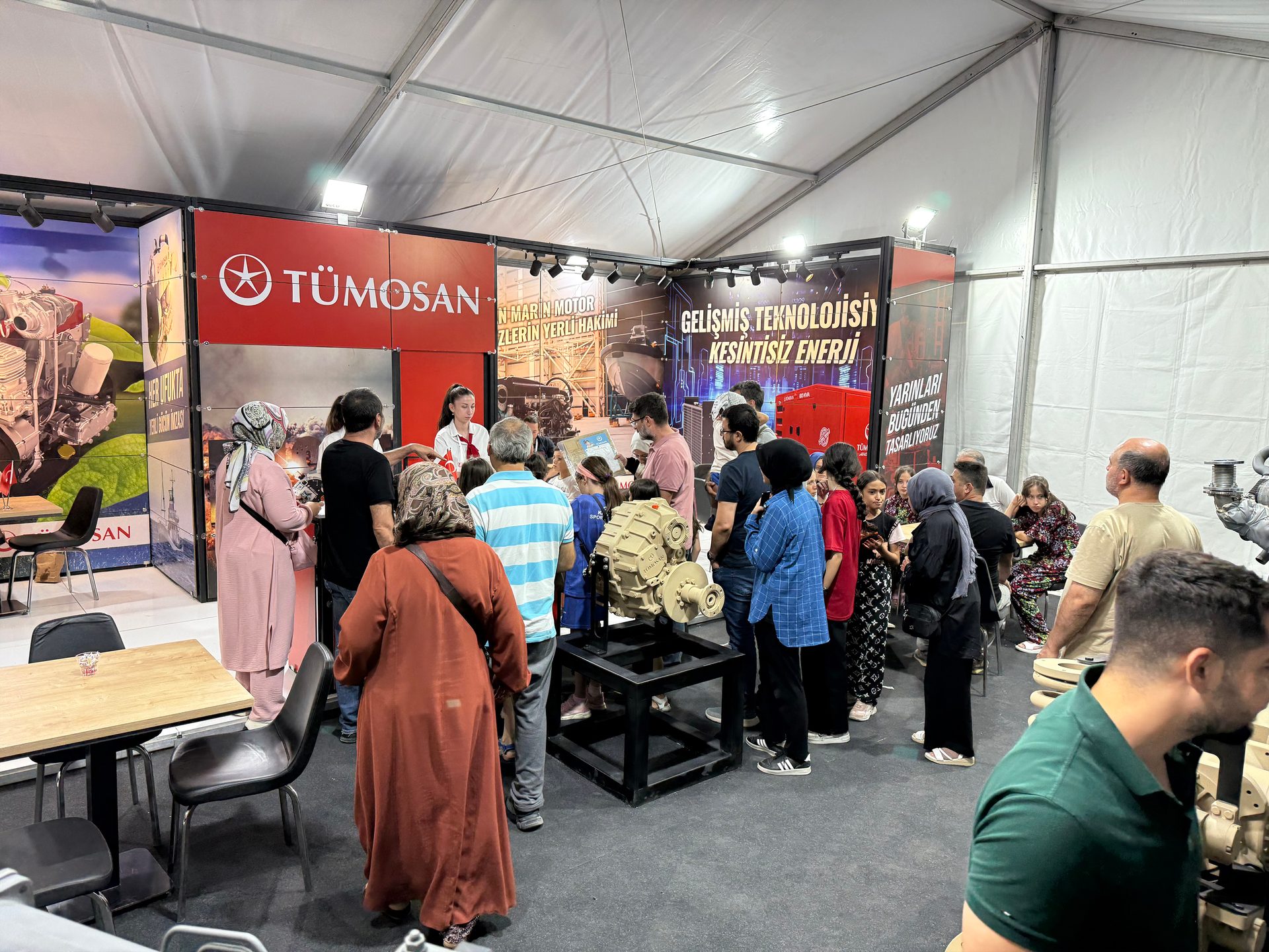 People at a TÜMOSAN exhibition booth under a tent, viewing machinery and tech displays.