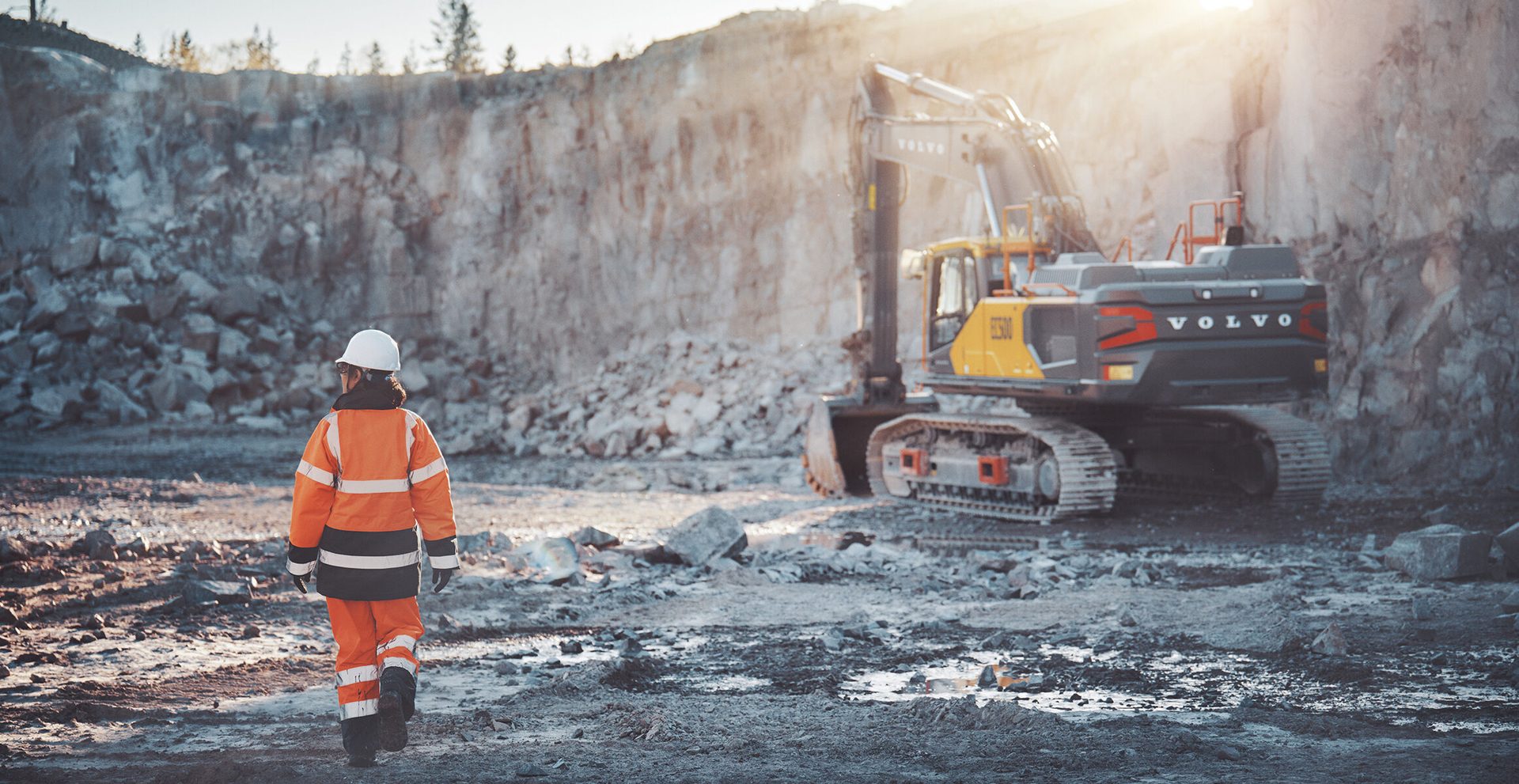 A construction worker approaches a large Volvo excavator in a sunny quarry.