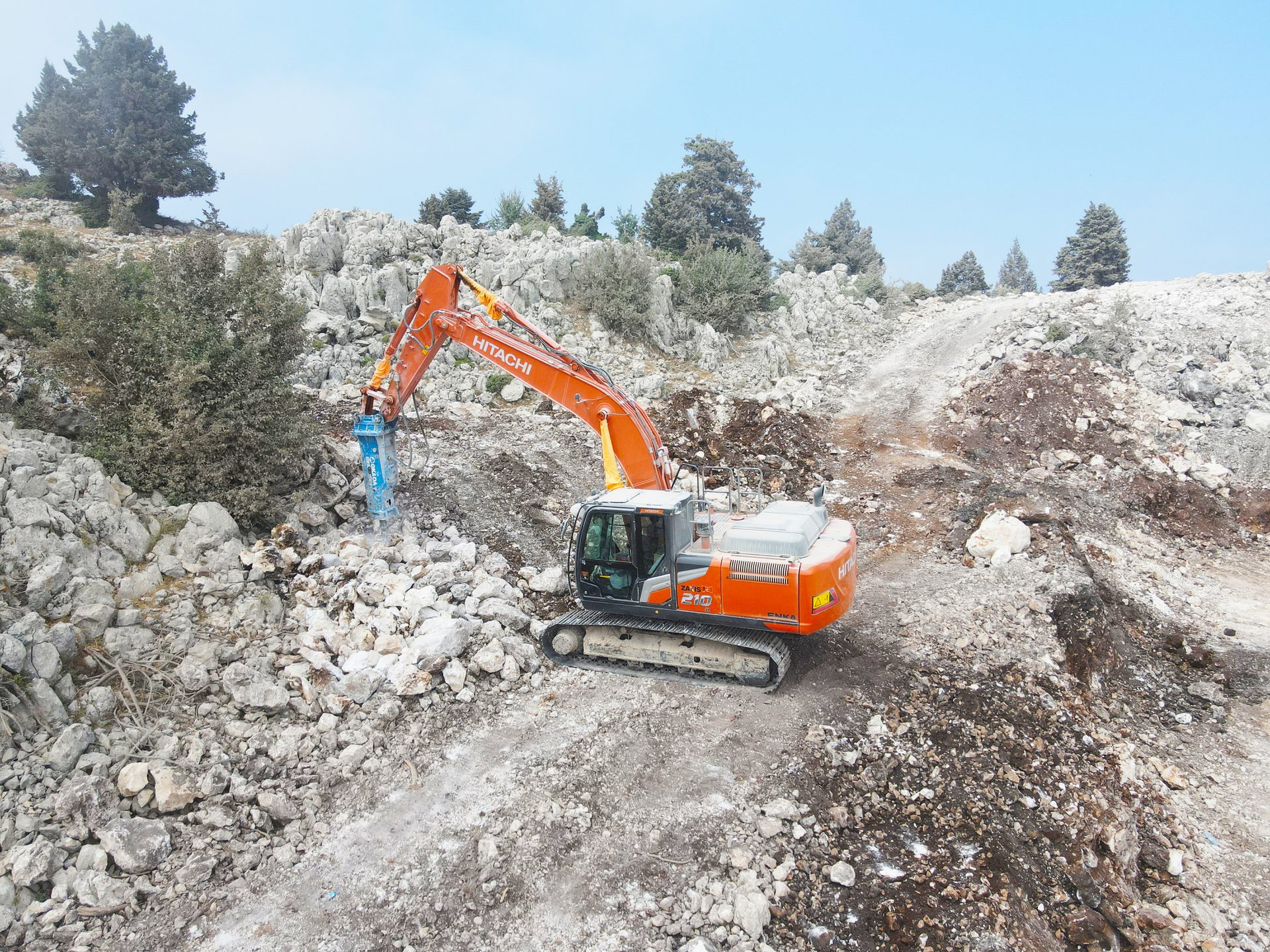 An orange Hitachi excavator with a hydraulic breaker attachment breaking rocks in a rocky landscape.