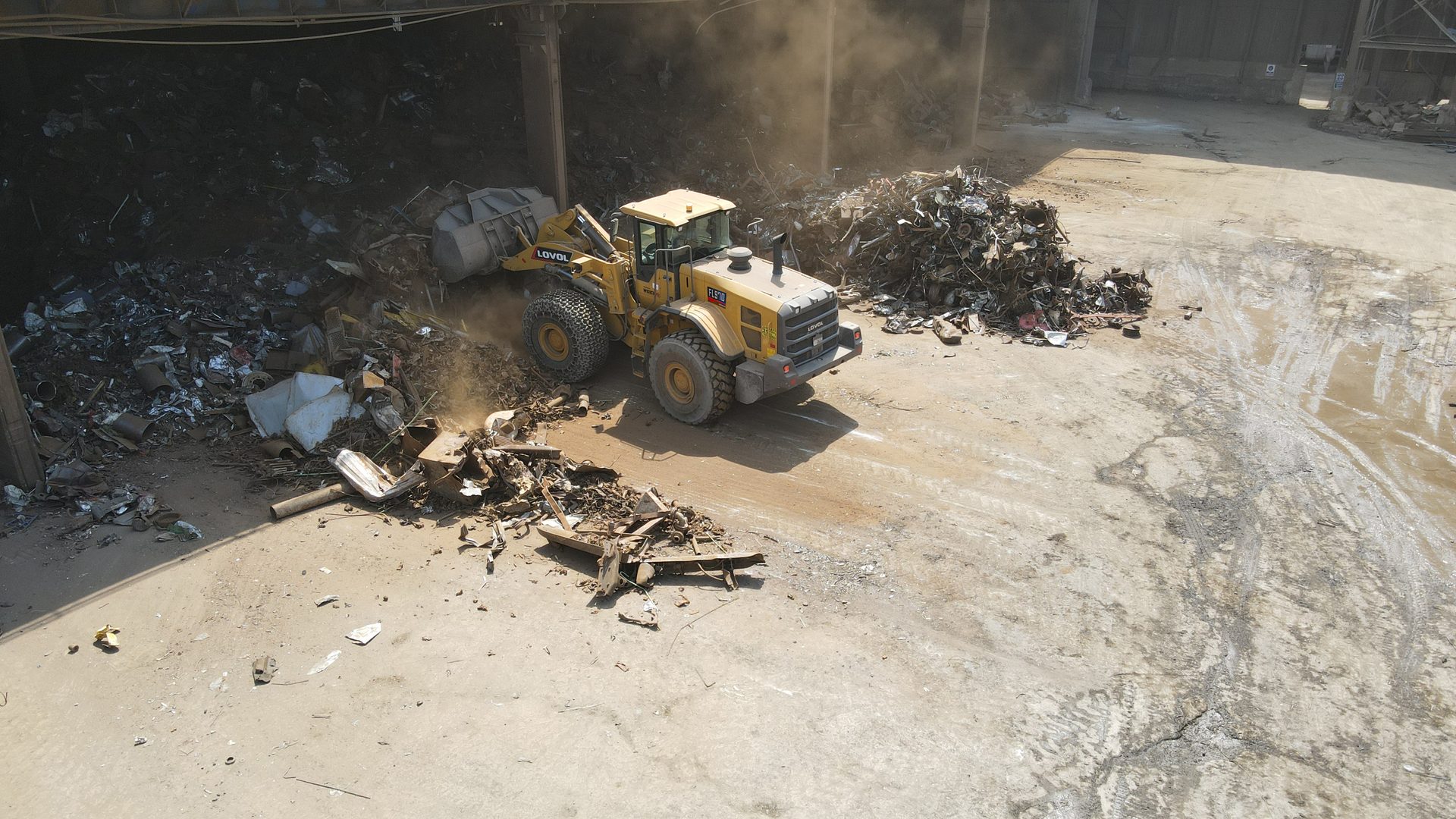 Yellow front loader moves scrap metal, kicking up dust, in a scrap yard.