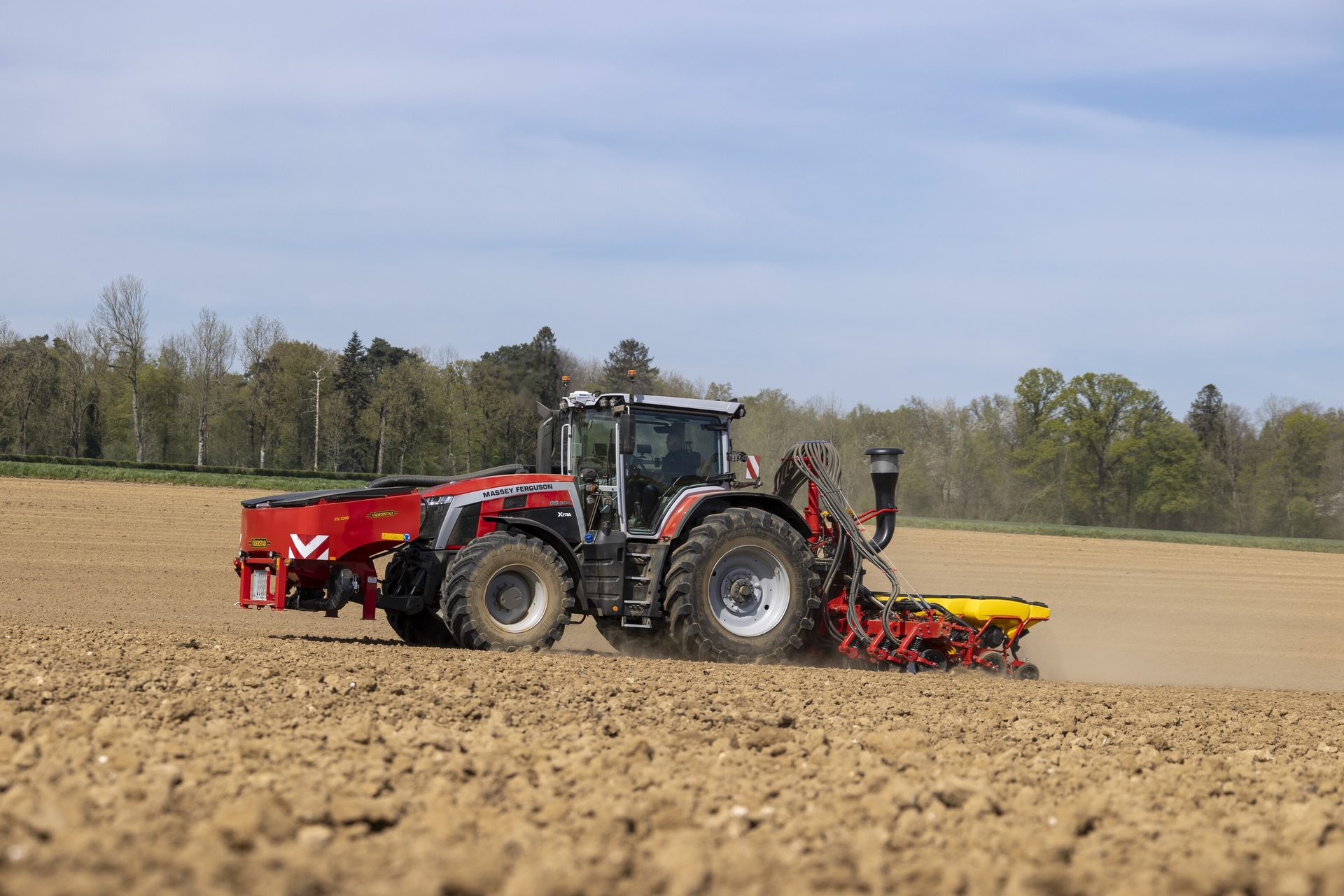 A red Massey Ferguson tractor with a seeder works a field under a partly cloudy sky.