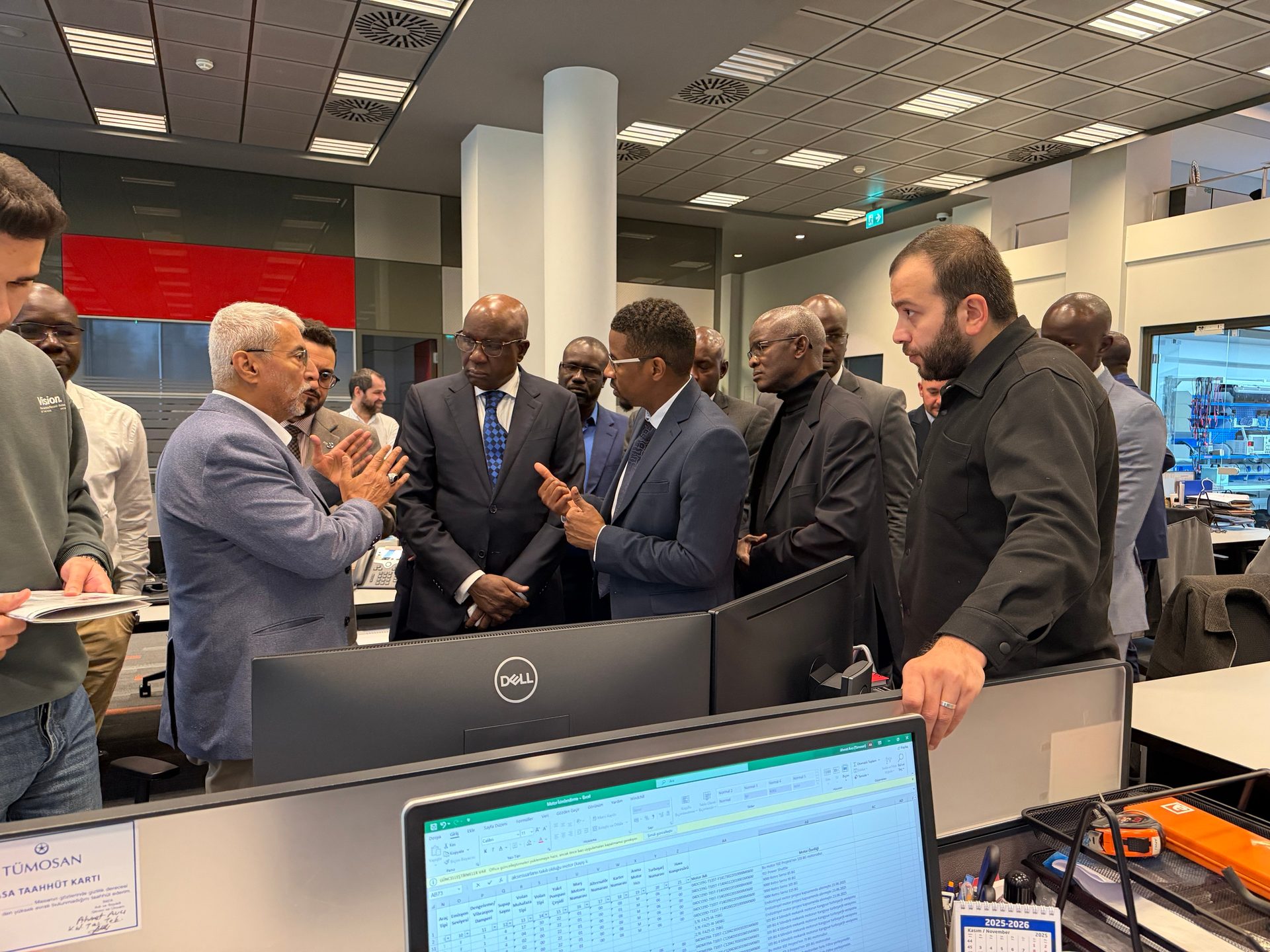 A group of men in an office, one speaking and gesturing near computer monitors with data.