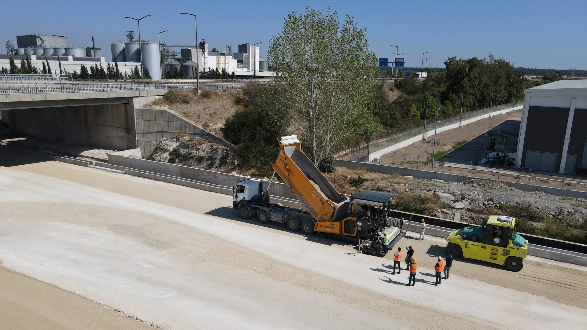 Aerial view of road construction: a paver lays asphalt from a dump truck, followed by a roller.