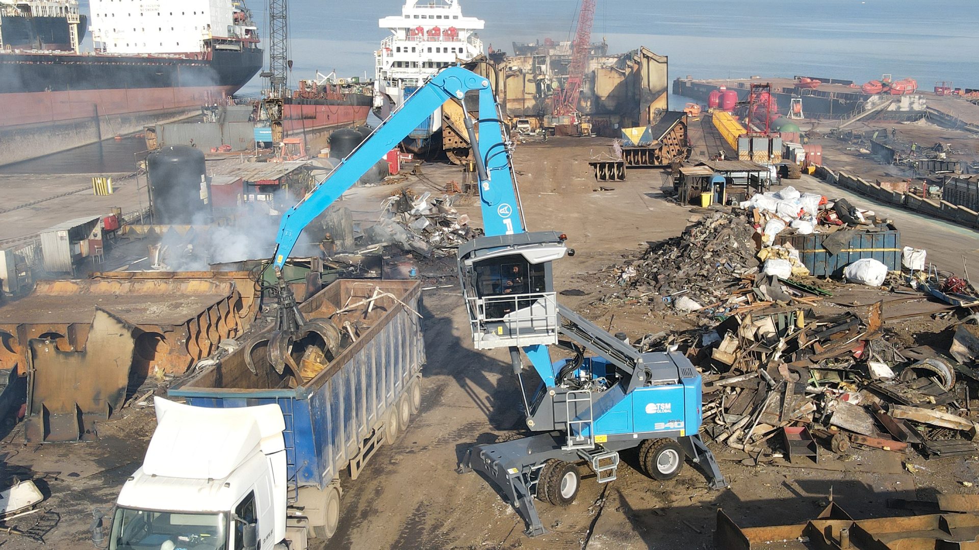A large blue material handler loads scrap metal into a truck at a busy industrial shipyard.