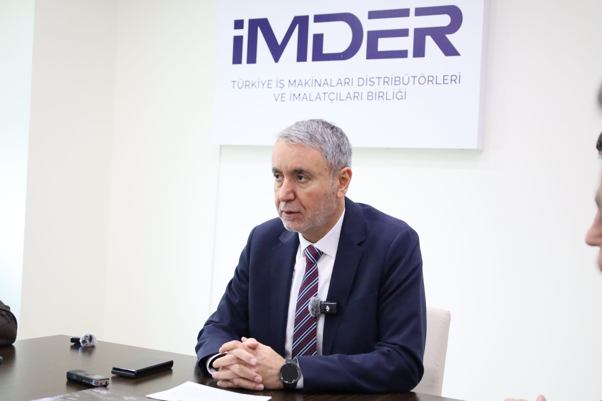 A man in a suit and tie sitting at a table with the iMDER logo in the background.