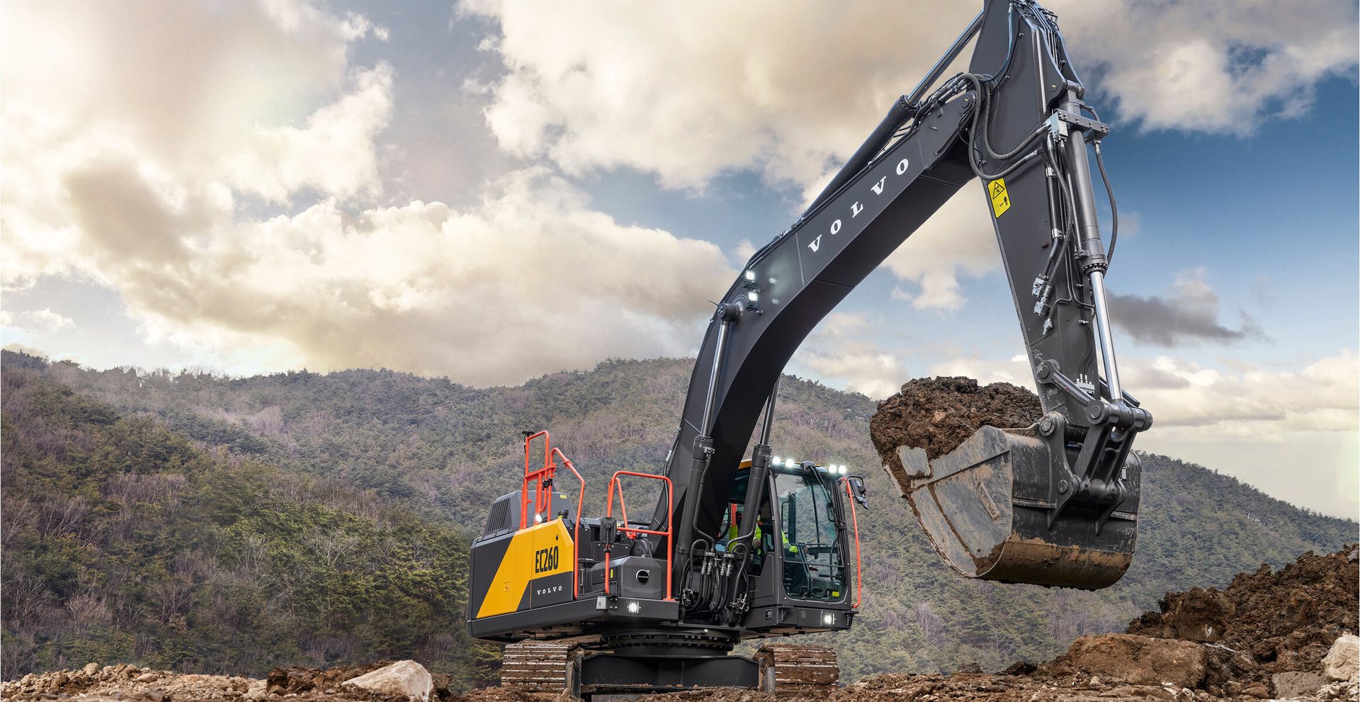 Volvo excavator with dirt in bucket, working in hilly terrain under cloudy sky.