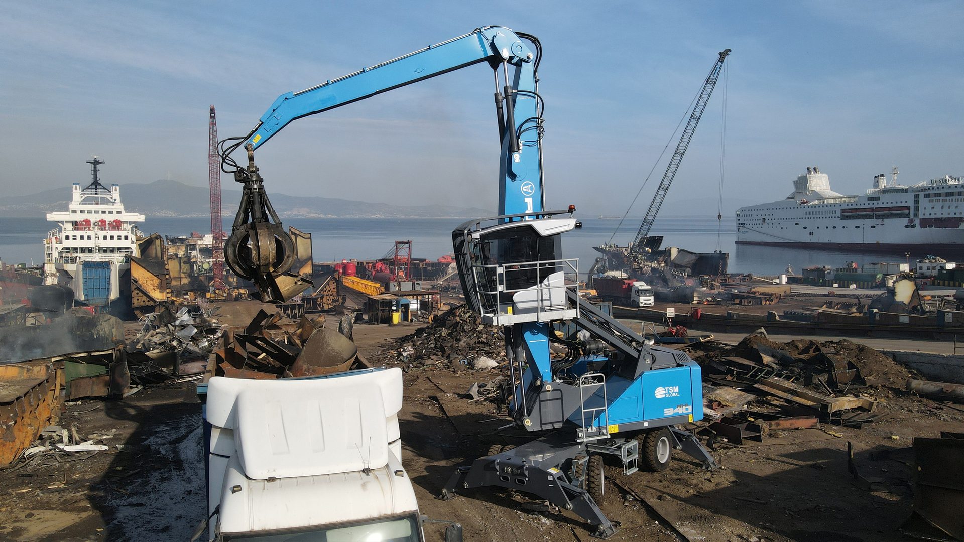 Blue material handler crane with a grapple loading scrap metal onto a white truck at a shipbreaking yard.