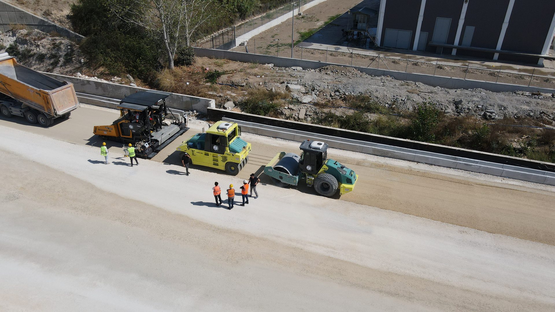 Aerial view of road construction with a dump truck, paver, two rollers, and workers.