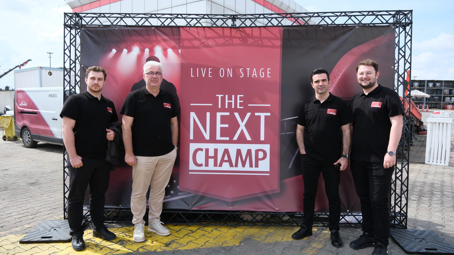 Four men in black shirts stand in front of a banner reading "THE NEXT CHAMP" outdoors.