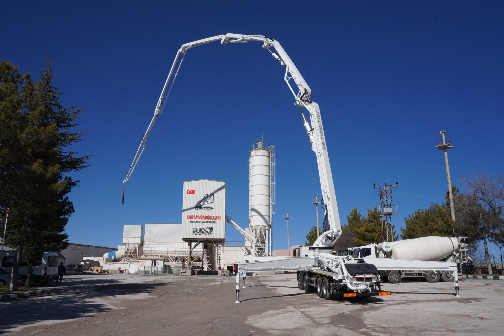 A large white concrete pump truck with extended boom, a concrete plant, and mixer truck under clear sky.