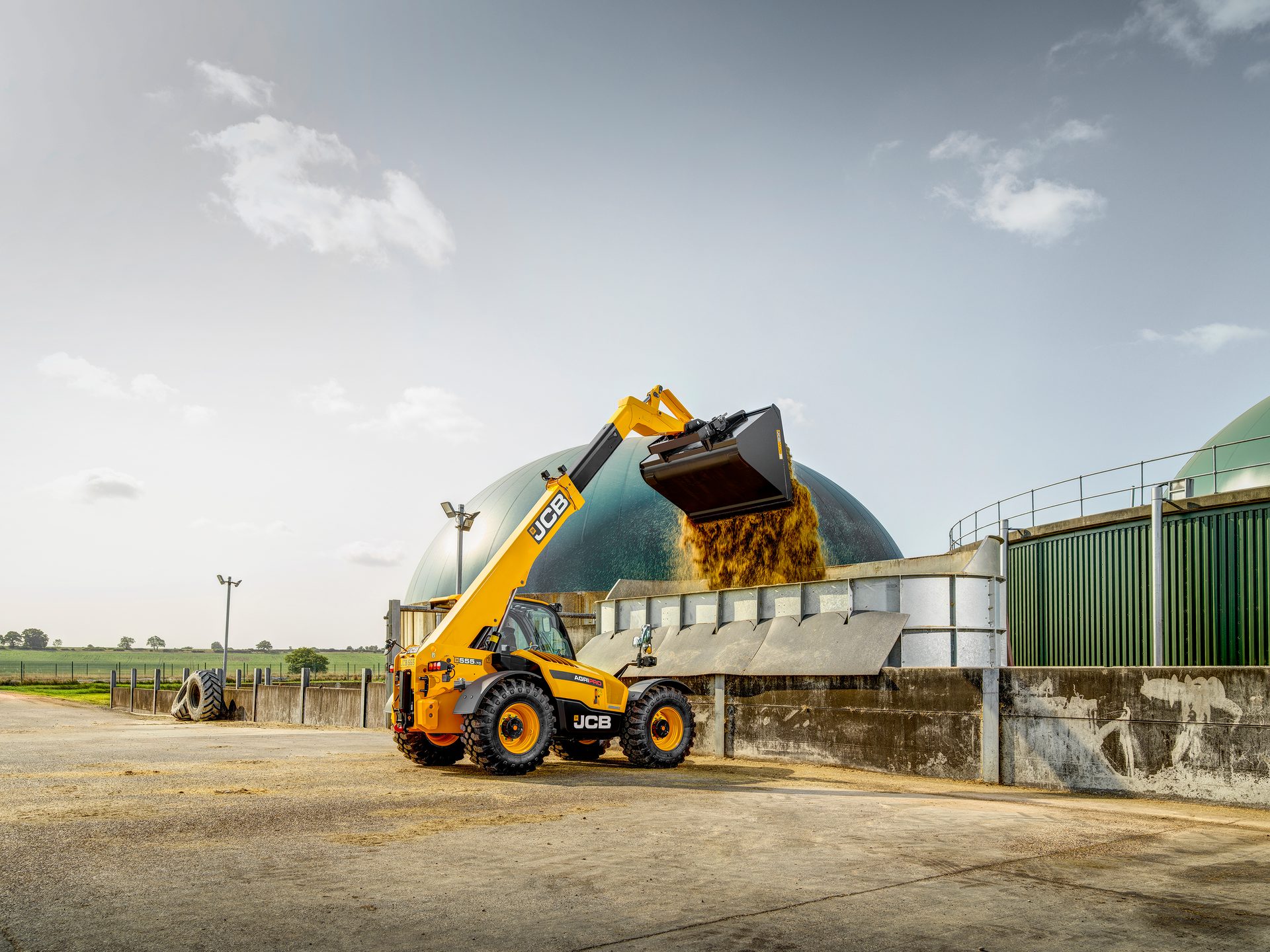Yellow JCB telehandler unloads organic material into a green domed biogas plant.
