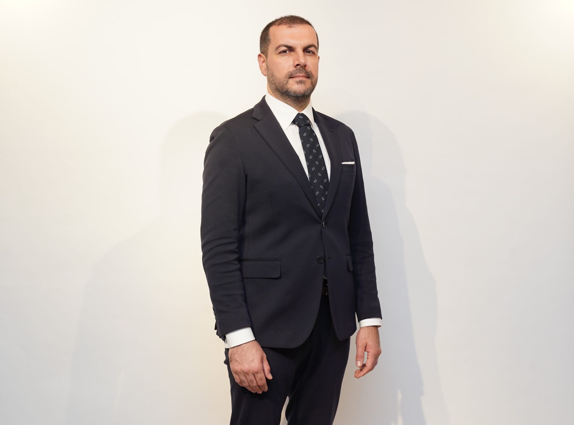 A man in a dark suit and tie stands against a white background.