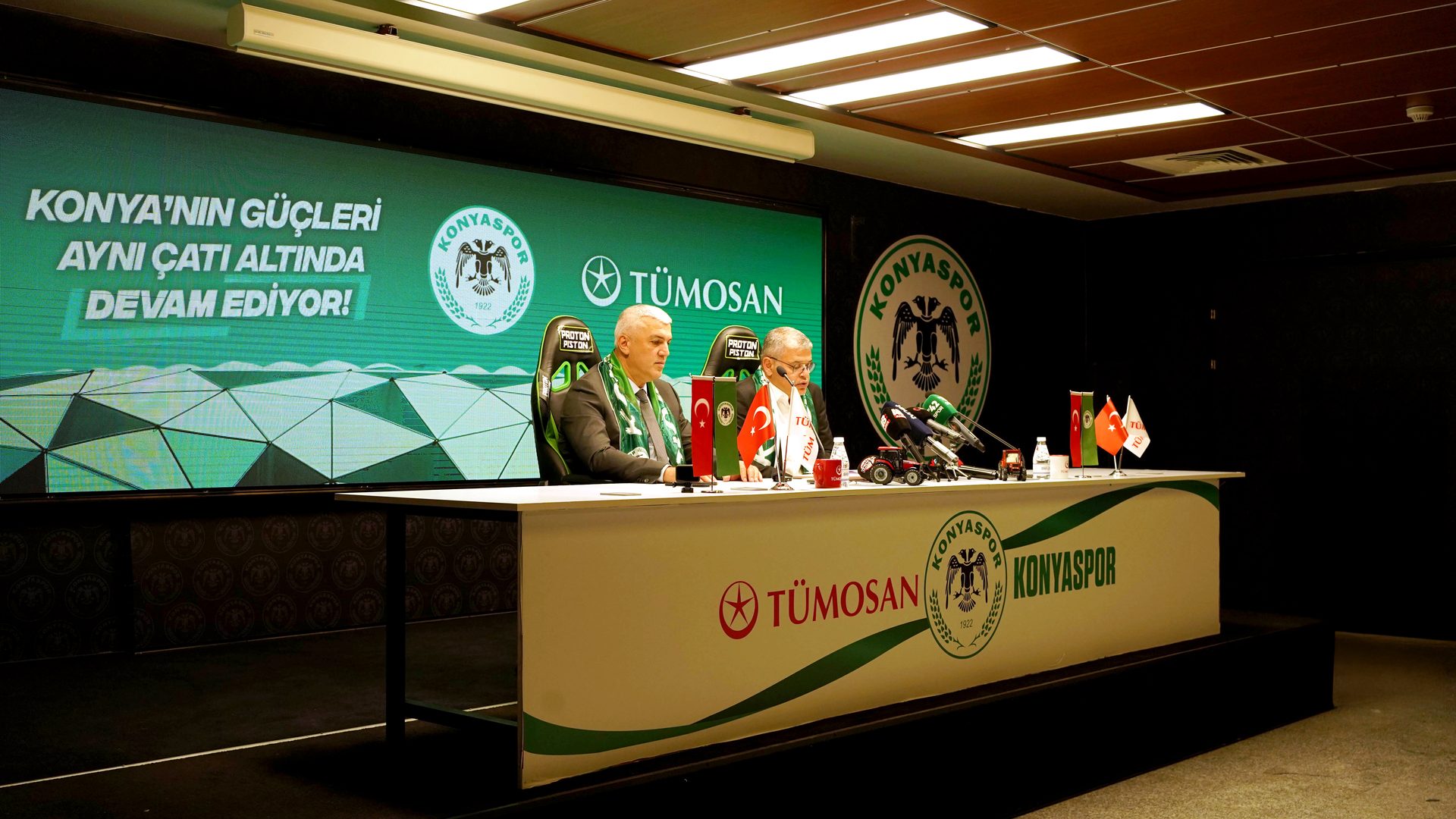 Press conference with two men, Konyaspor & Tümosan branding, flags, and a screen.