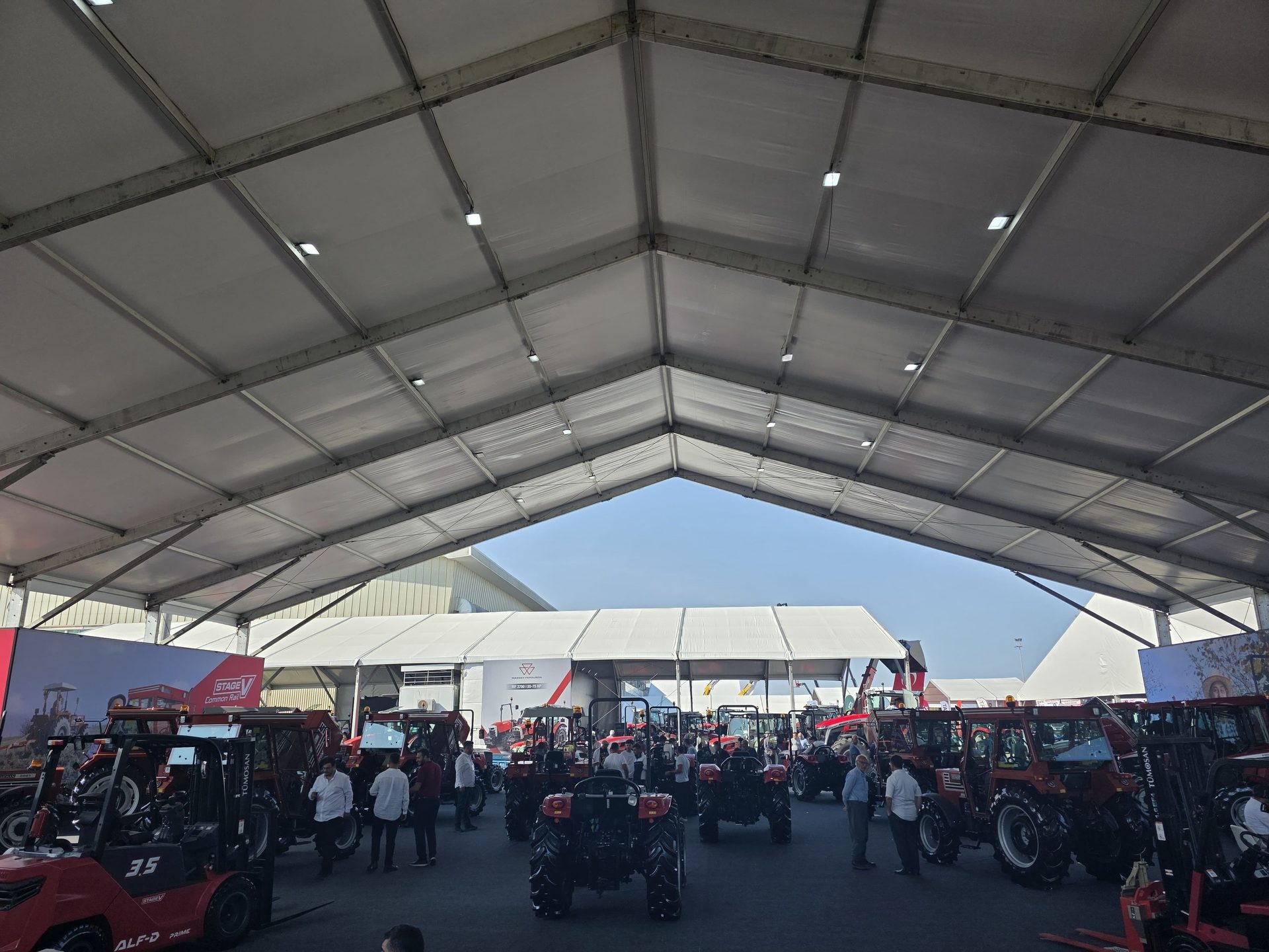 People view tractors and forklifts at a machinery exhibition inside a large tent.