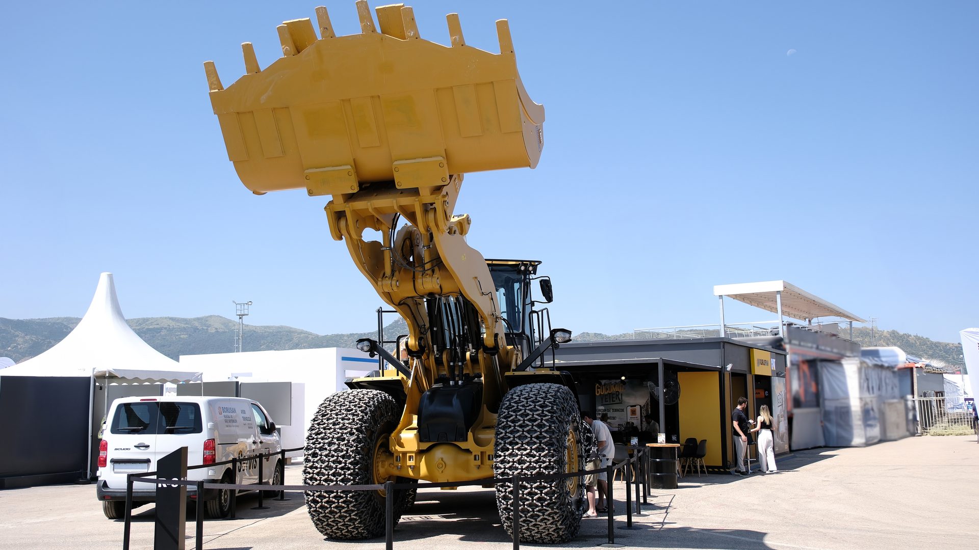 A large yellow wheel loader with a raised bucket at an outdoor event under a clear sky.
